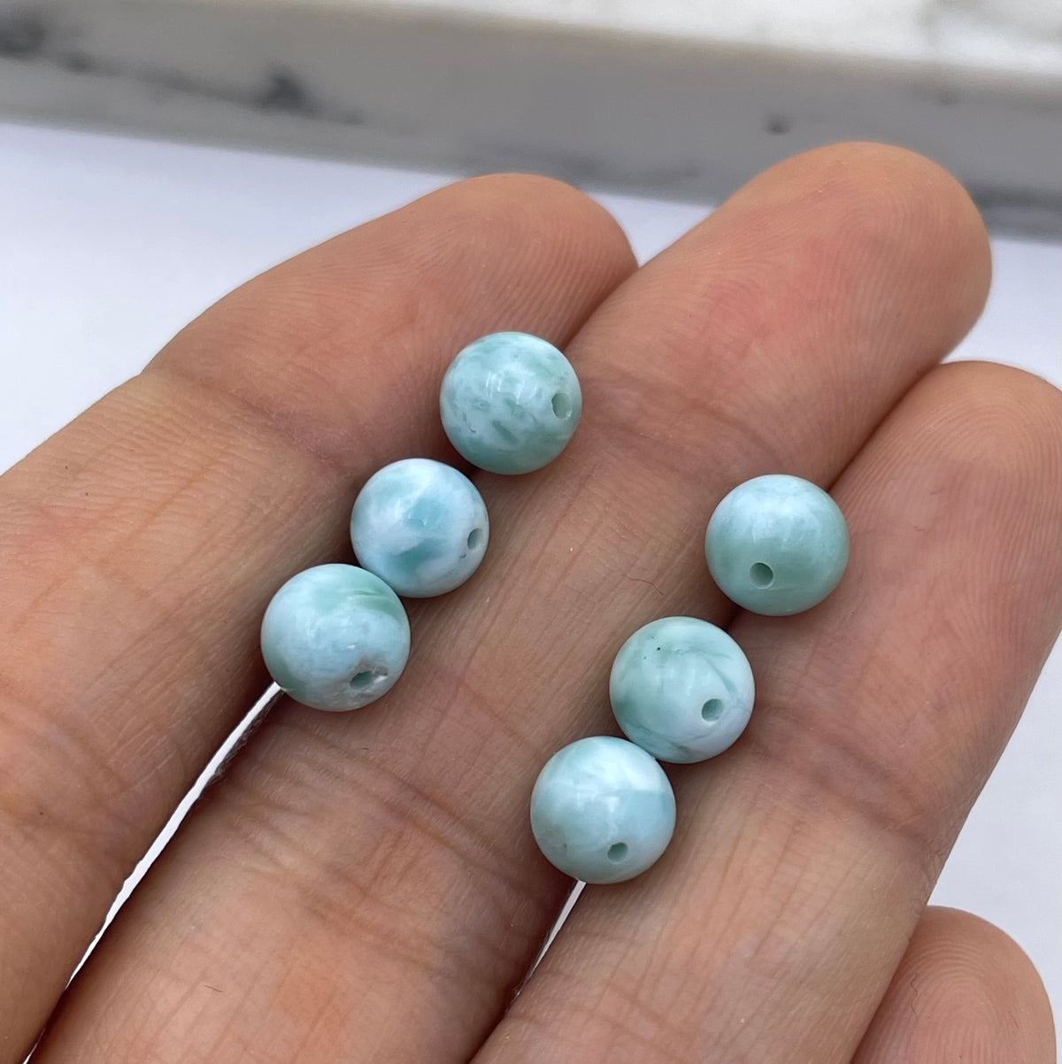 Six light blue larimar round beads with a hole in the center, displayed on a person's fingers against a white background.