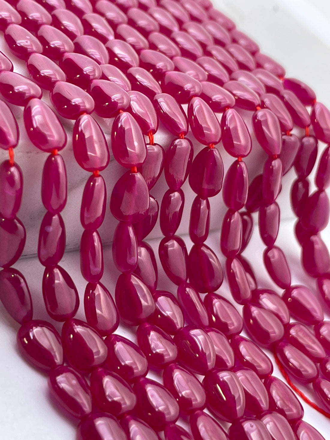 Close-up of glossy, teardrop-shaped lab ruby beads strung in multiple strands on a white surface, showing a vibrant magenta-pink color.
