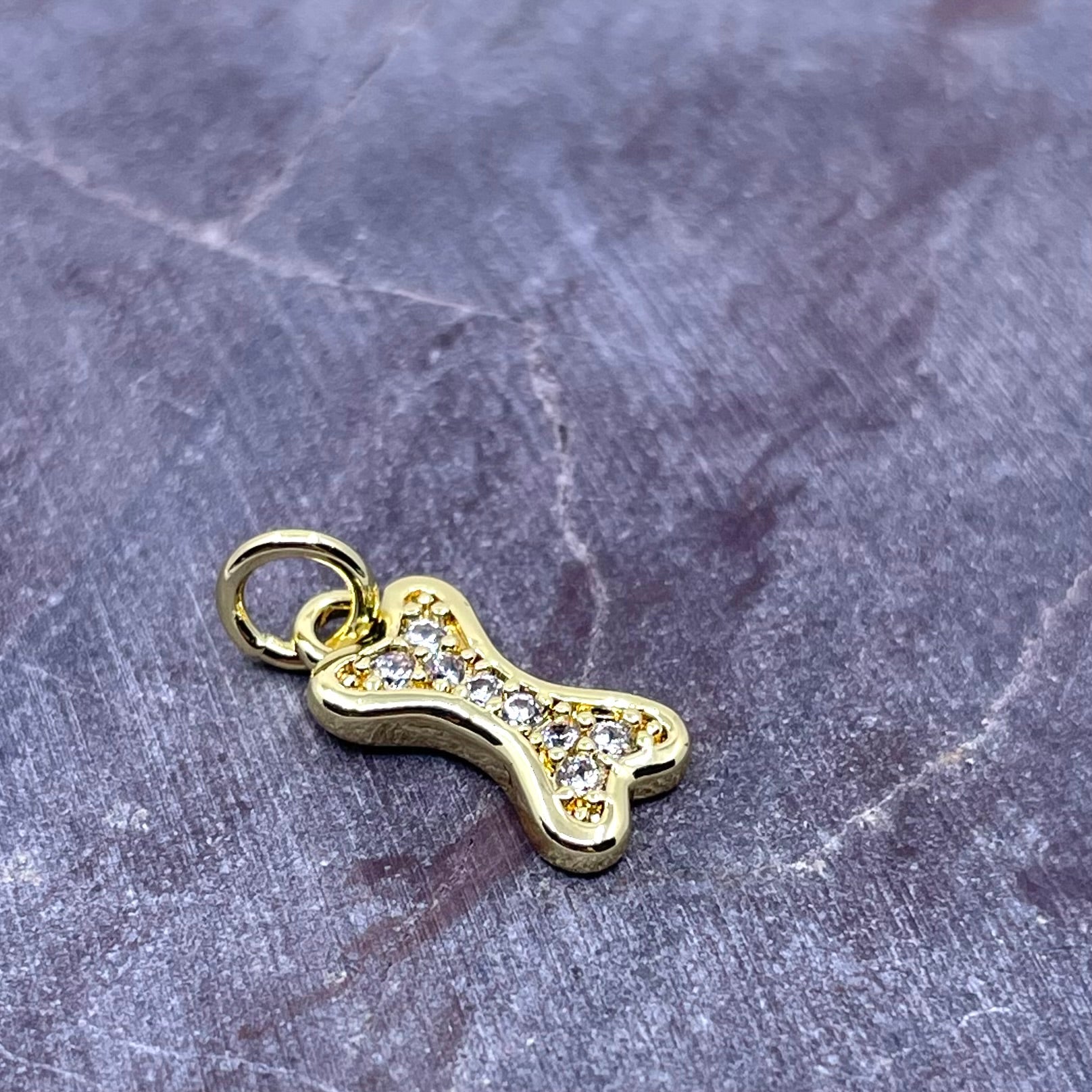 A gold bone-shaped charm or pendant embellished with small clear stones, resting on a textured gray stone surface.
