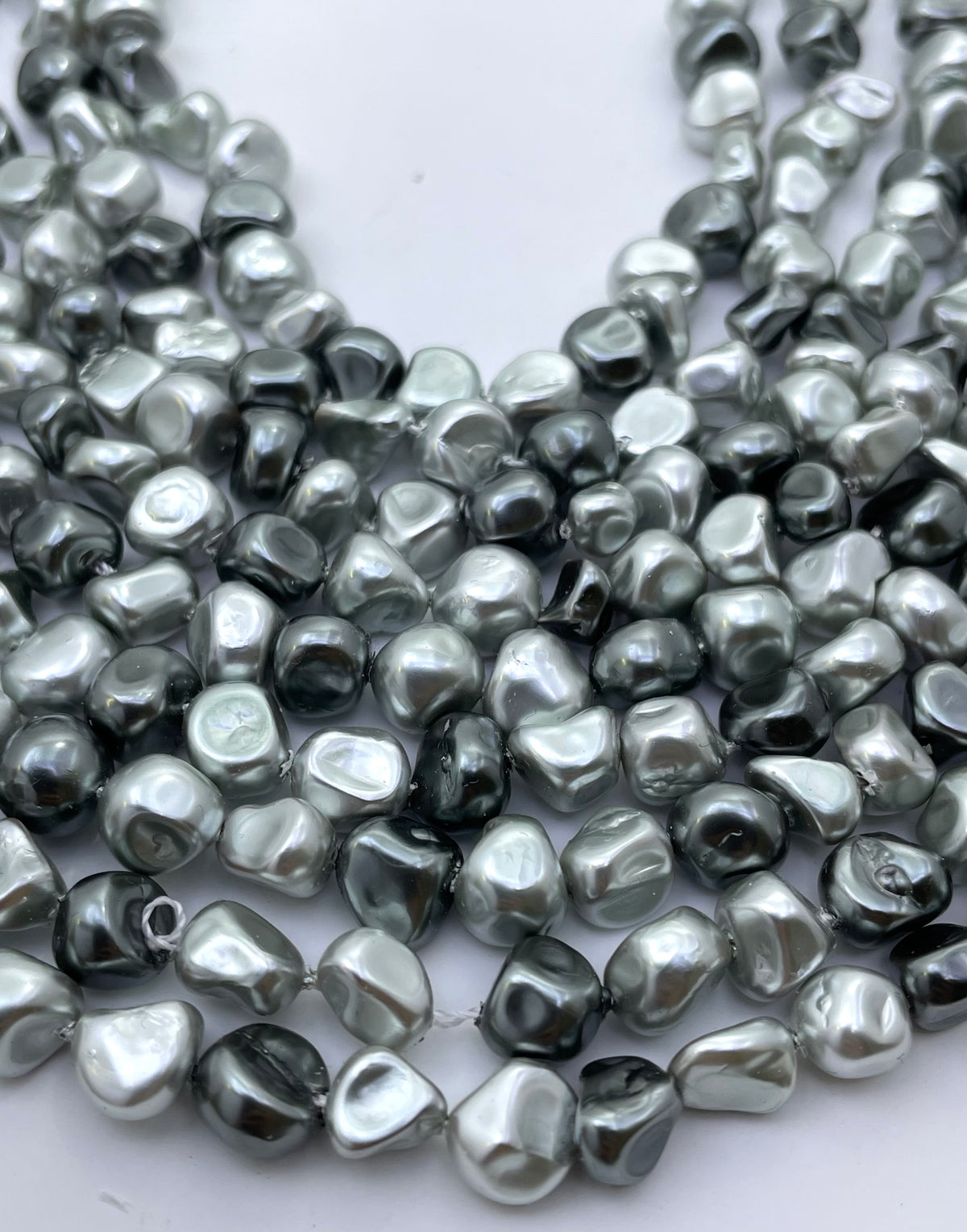 Close-up of multiple strands of irregular, polished shell pearl beads in a metallic silver-gray color, arranged on a plain white background.