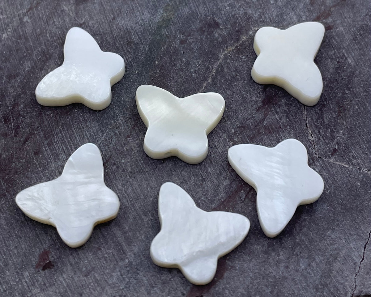 Six white mother-of-pearl butterfly-shaped beads arranged on a dark textured slate surface.