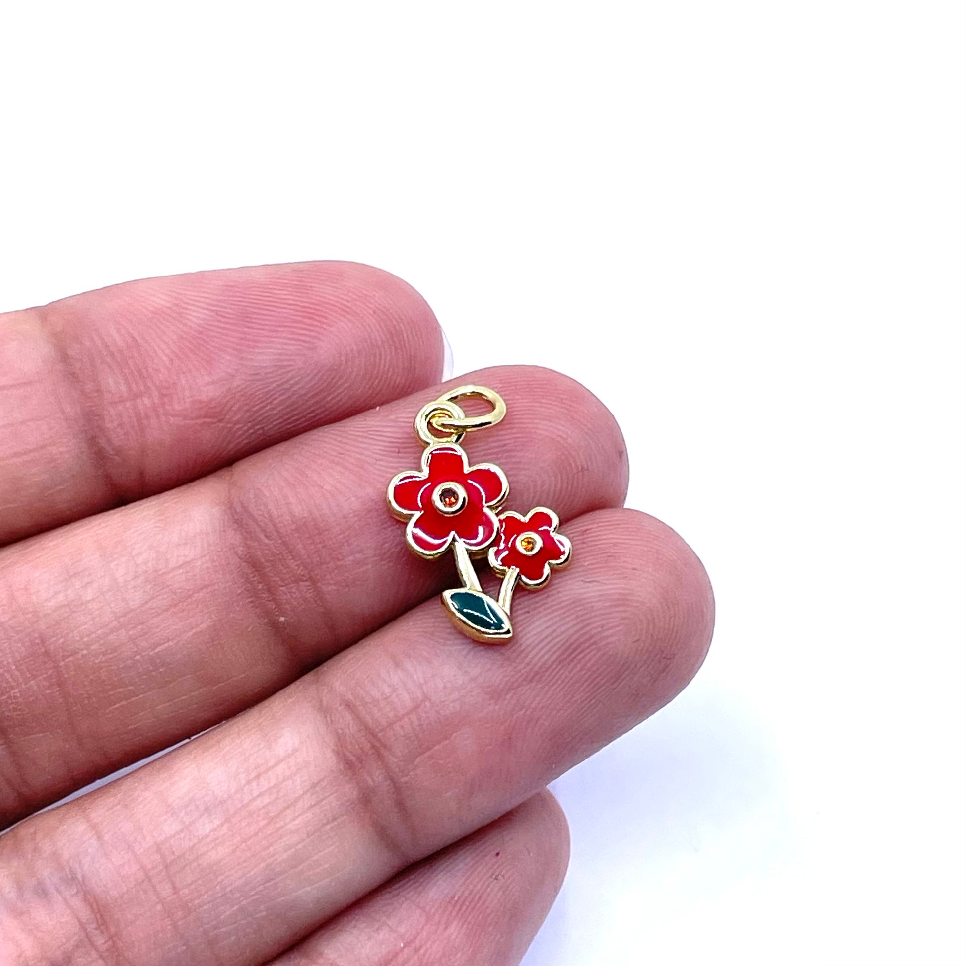 A small gold-tone charm shaped like two red flowers with green leaves, held between a person's fingers against a white background.