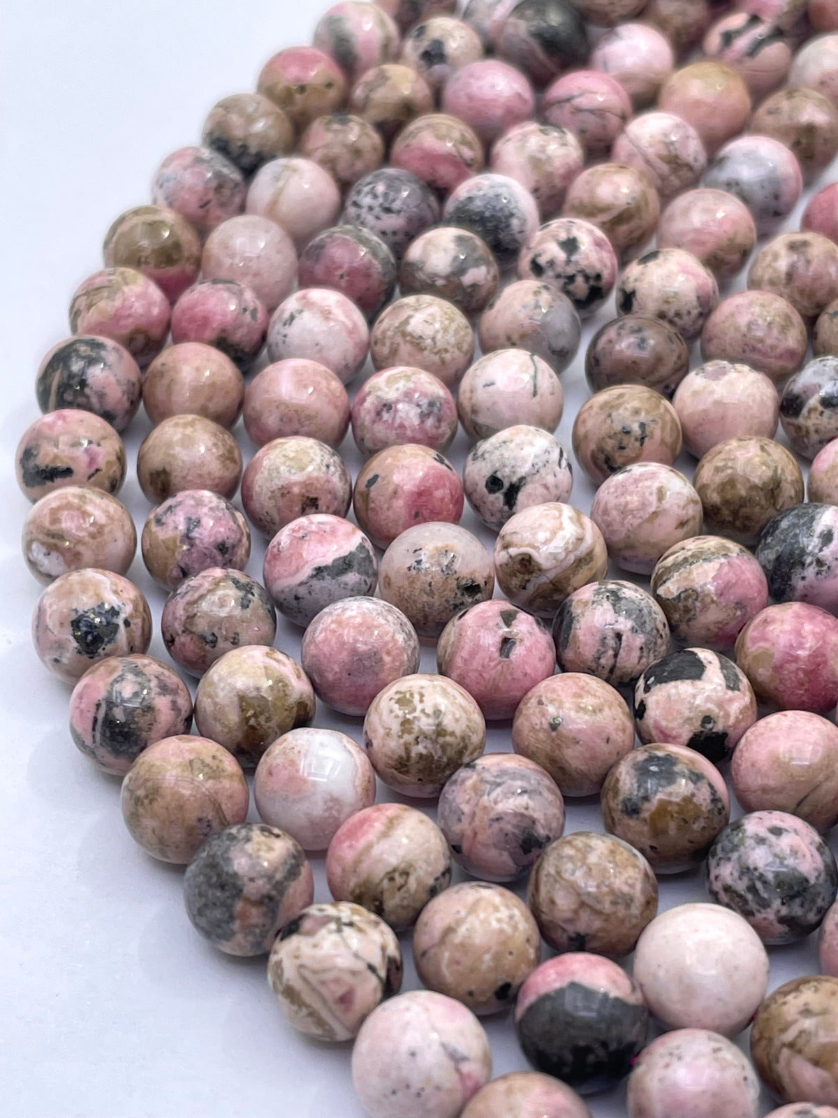 A close-up of a strand of round, polished pink and beige rhodochrosite stone beads with black and white marbled patterns, arranged on a white surface.