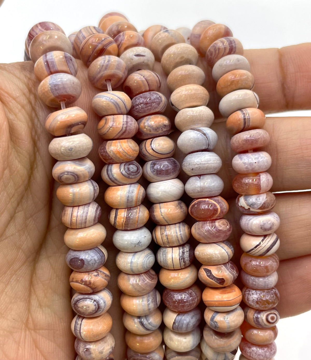 Multiple strands of polished gobi agate stone beads with natural swirling patterns in beige, brown, orange, and purple, held in a person's hand against a white background.