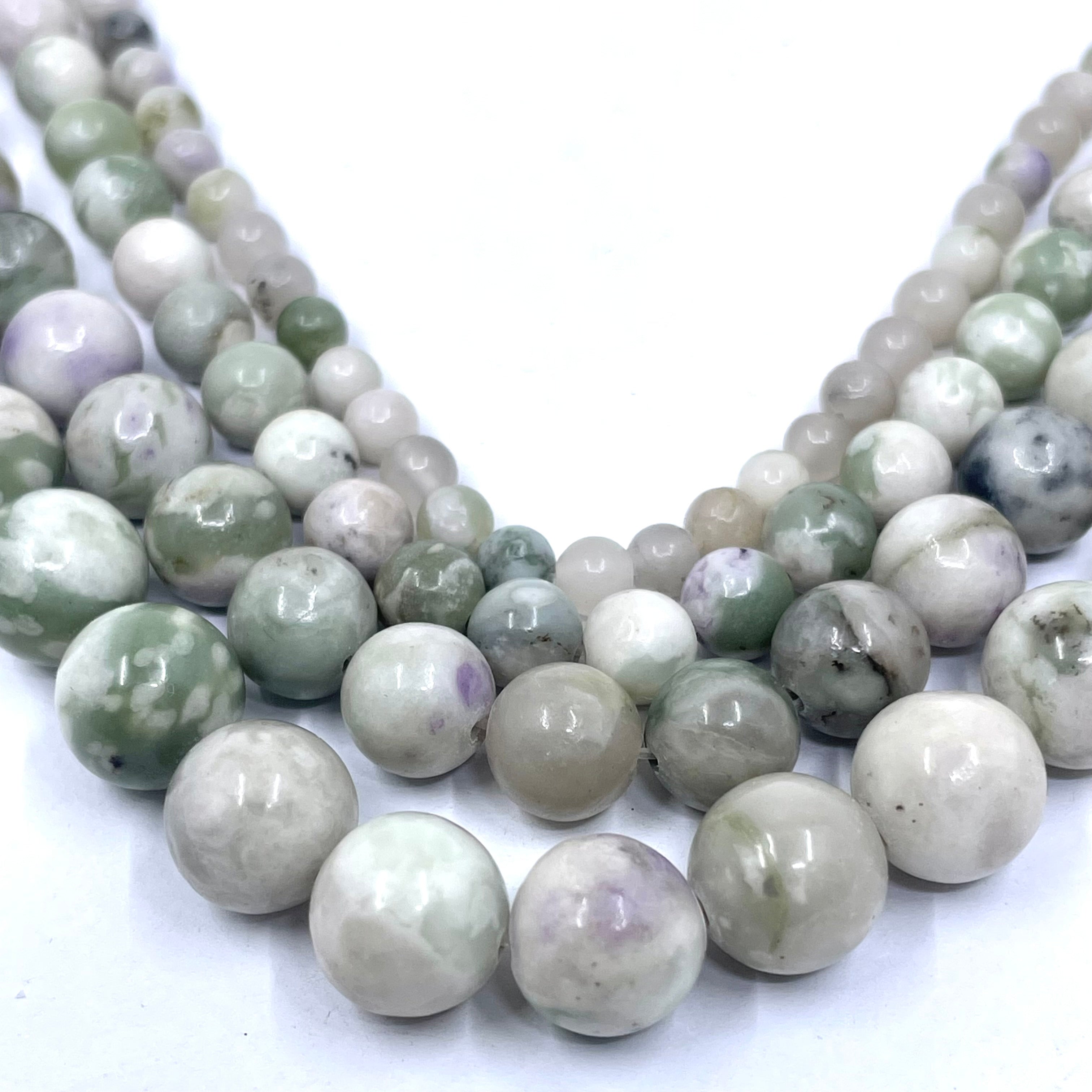 Multiple strands of polished round peace jade gemstone beads in shades of pale green, gray, white, and soft lavender, arranged in a V shape on a white background.