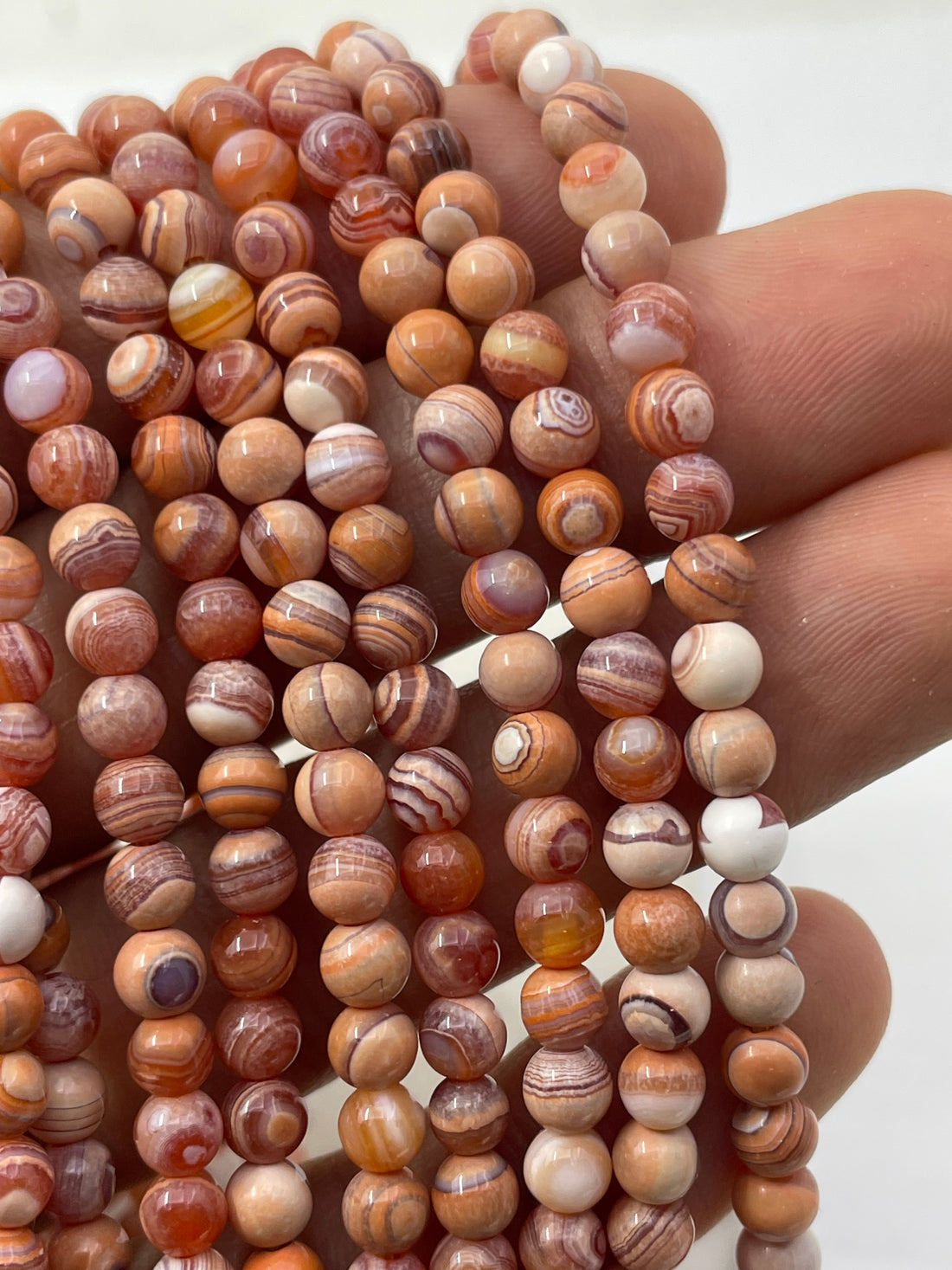 A close-up of polished round gobi agate gemstone beads in earthy orange, peach, cream, and lavender-brown banding patterns, arranged on a plain white background.
