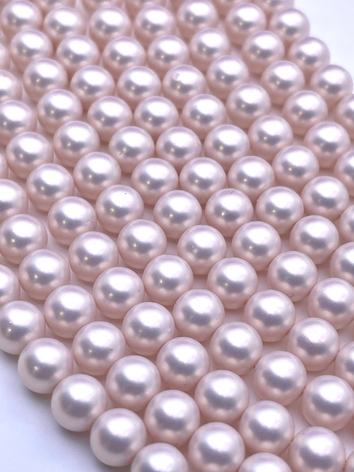 A close-up of multiple strands of round, shell pearls in a soft blush pink color, arranged diagonally on a plain white background.
