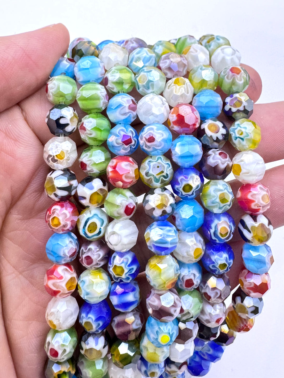 A hand holding a bundle of colorful glass lampwork beads with floral and marbled patterns in shades of blue, green, white, red, yellow, and black.