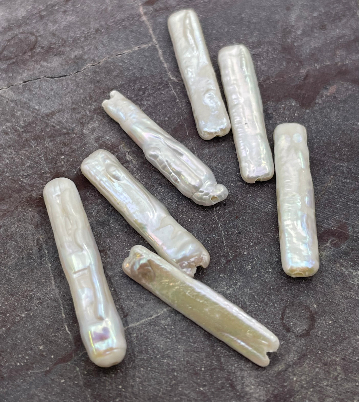Seven natural baroque freshwater pearl beads with an iridescent finish, displayed on a dark textured slate surface.