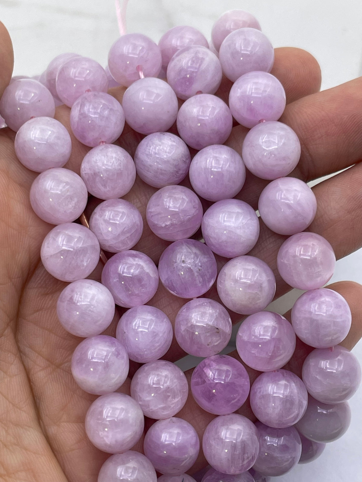 A hand holding a strand of polished round kunzite beads in light lavender purple, likely amethyst gemstone beads, against a light background.