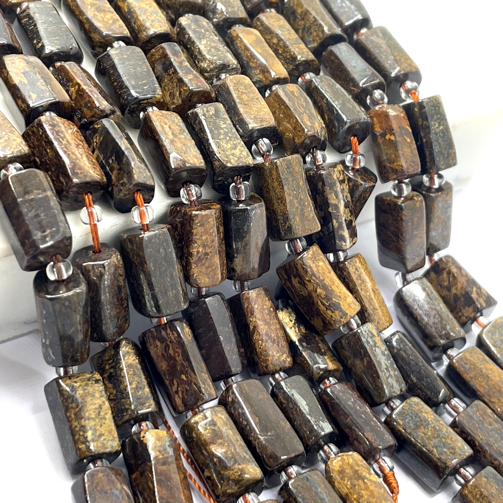 A close-up of a strand of polished rectangular bronzite beads made from dark brown and black stone with natural golden-brown veining, strung on a cord with small metallic spacer rings.
