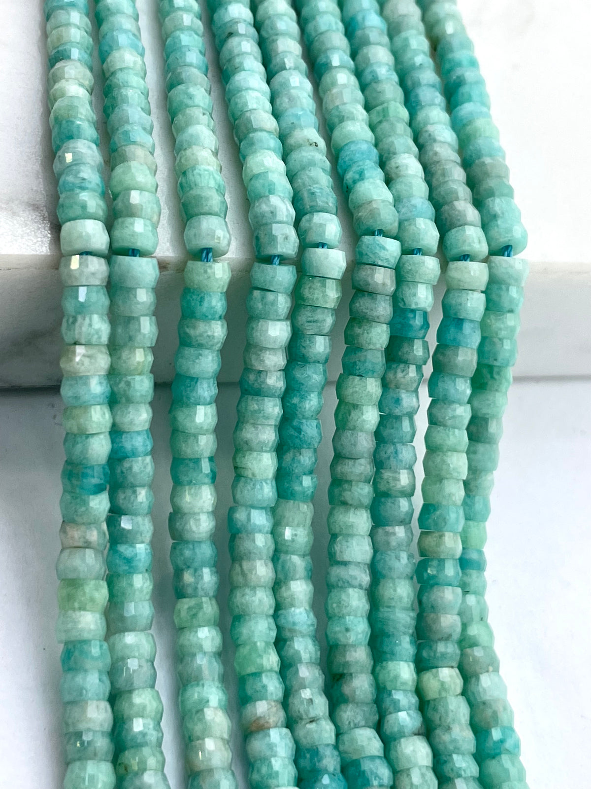 Multiple strands of faceted amazonite beads in a light aqua-green stone, arranged vertically on a white marble surface.