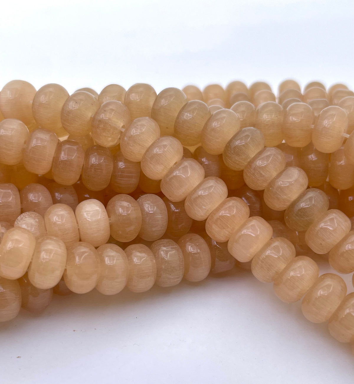 Close-up of multiple strands of small cats eye cylindrical beads in a warm translucent peachy-tan color on a white background.