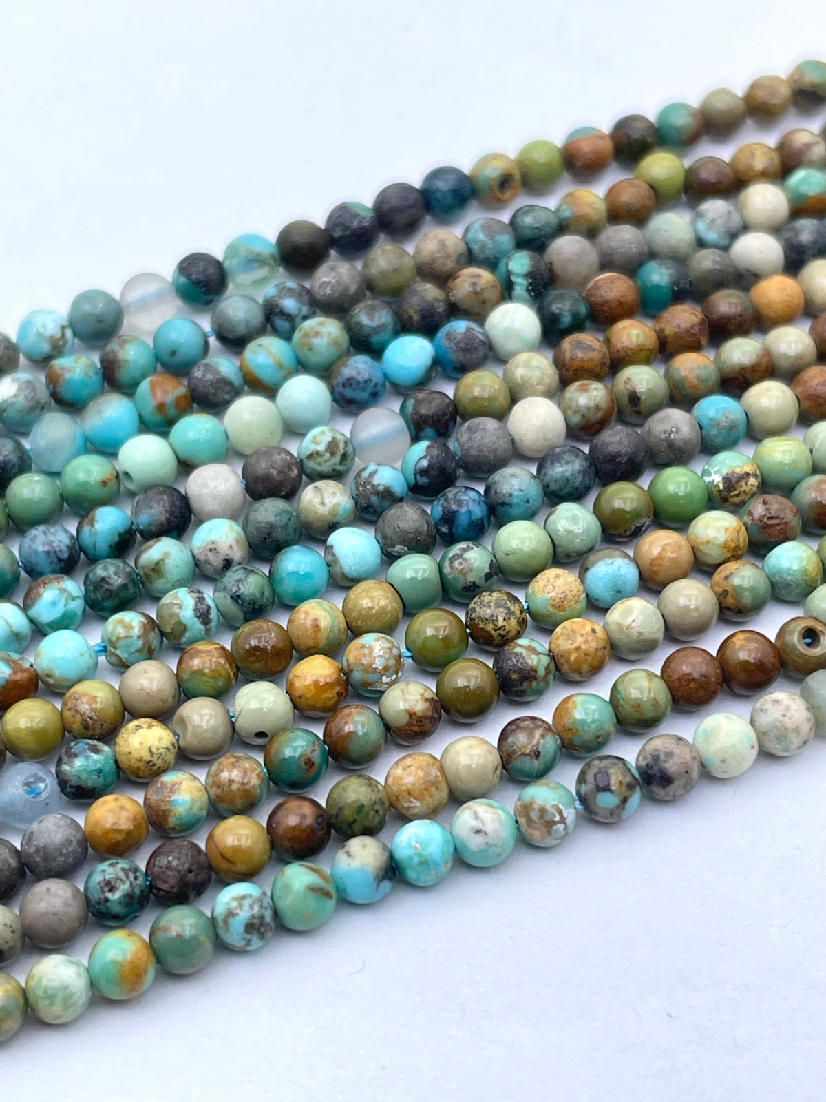 Multiple strands of round turquoise-stone beads with natural mottled patterns in blue, green, brown, gray, and cream, arranged diagonally on a plain white background.
