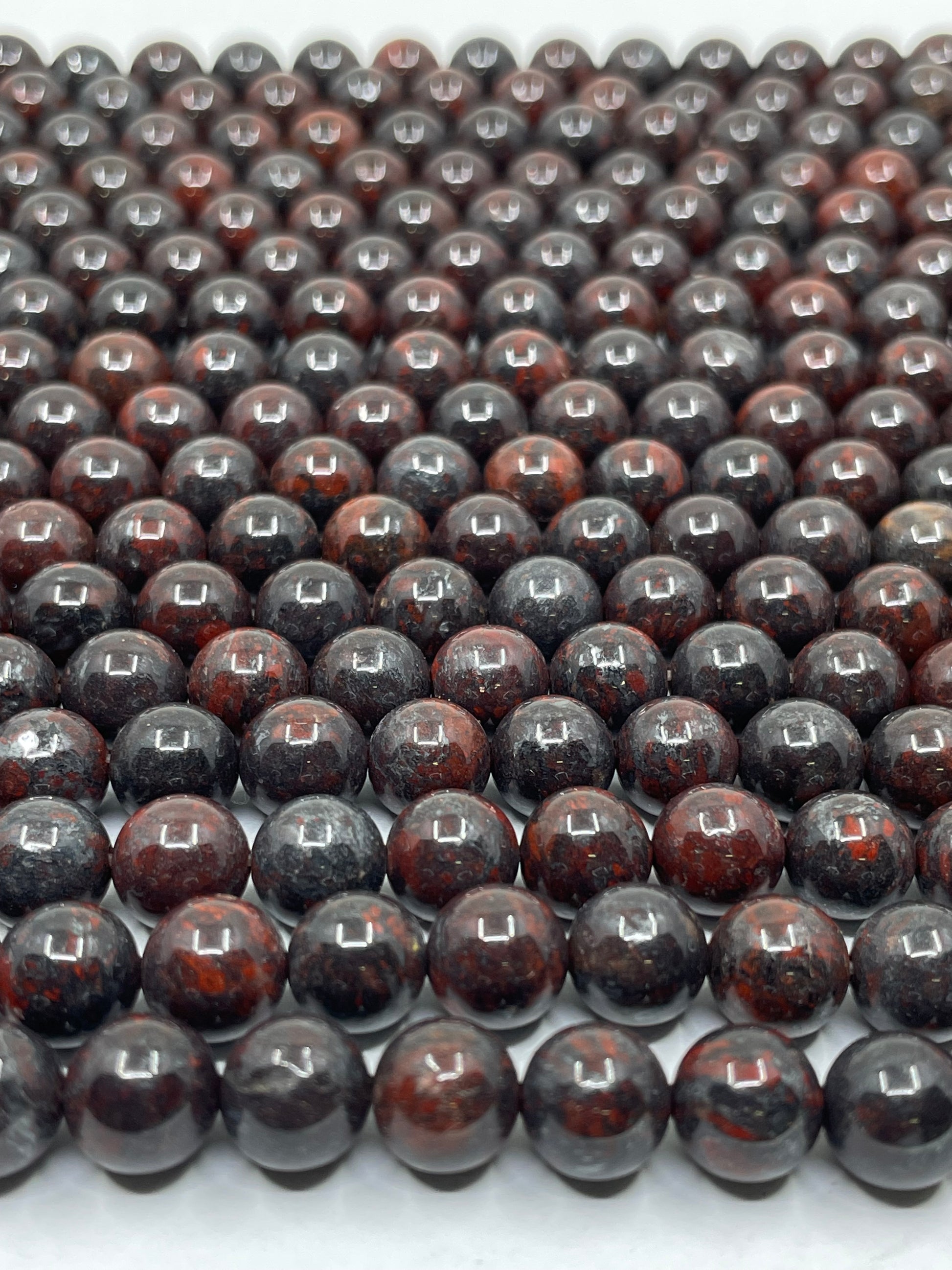 A close-up of multiple strands of polished round brecciated jasper beads, in a dark reddish-brown and black mottled pattern, arranged in rows on a white surface.