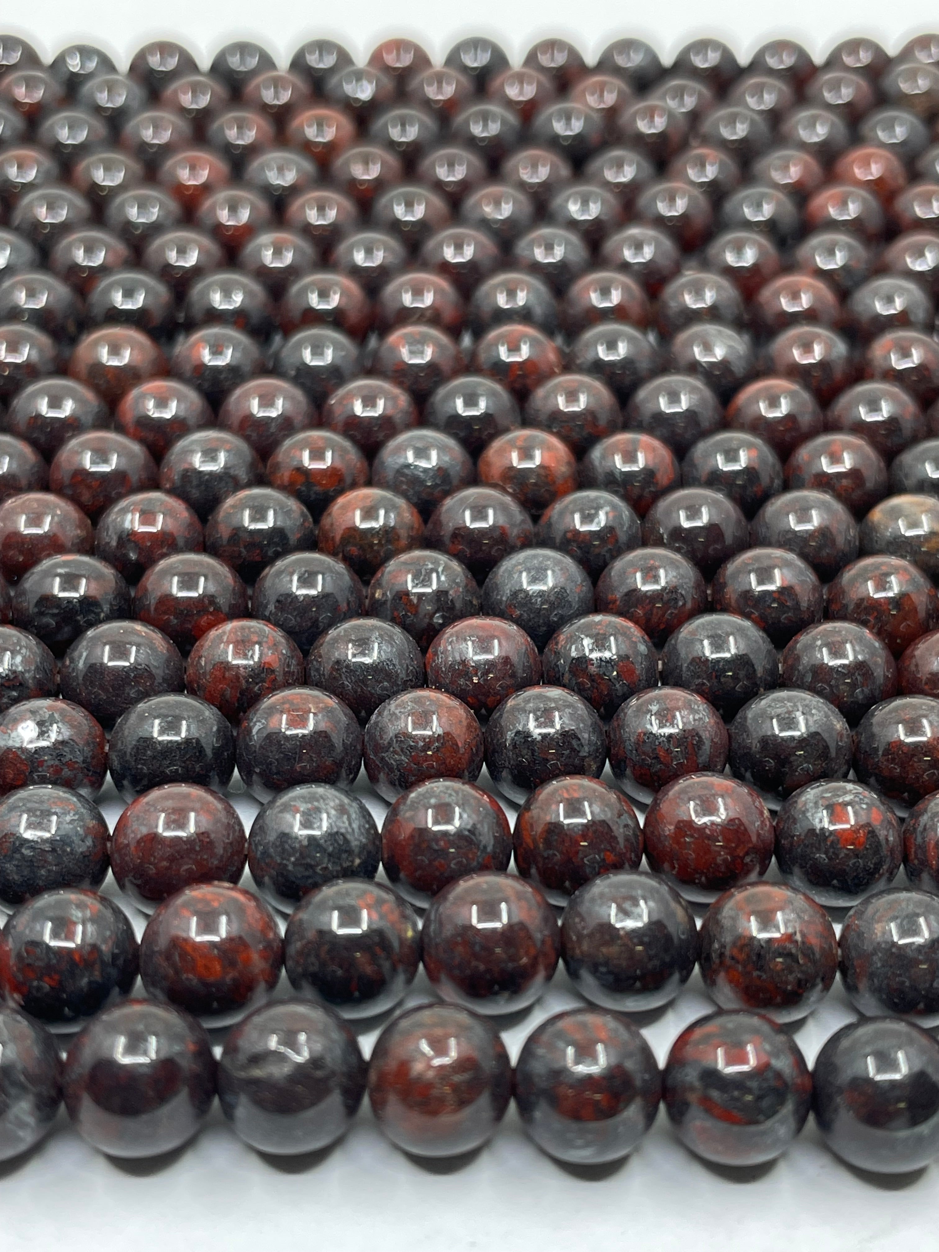 A close-up of multiple strands of polished round brecciated jasper beads, in a dark reddish-brown and black mottled pattern, arranged in rows on a white surface.