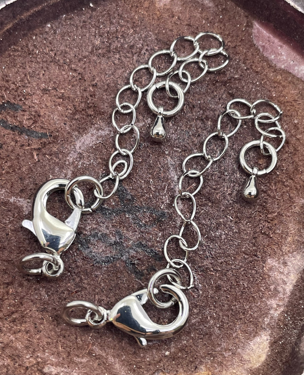 A silver-toned chain bracelet with lobster clasp ends and circular link details, laid on a textured brown surface.