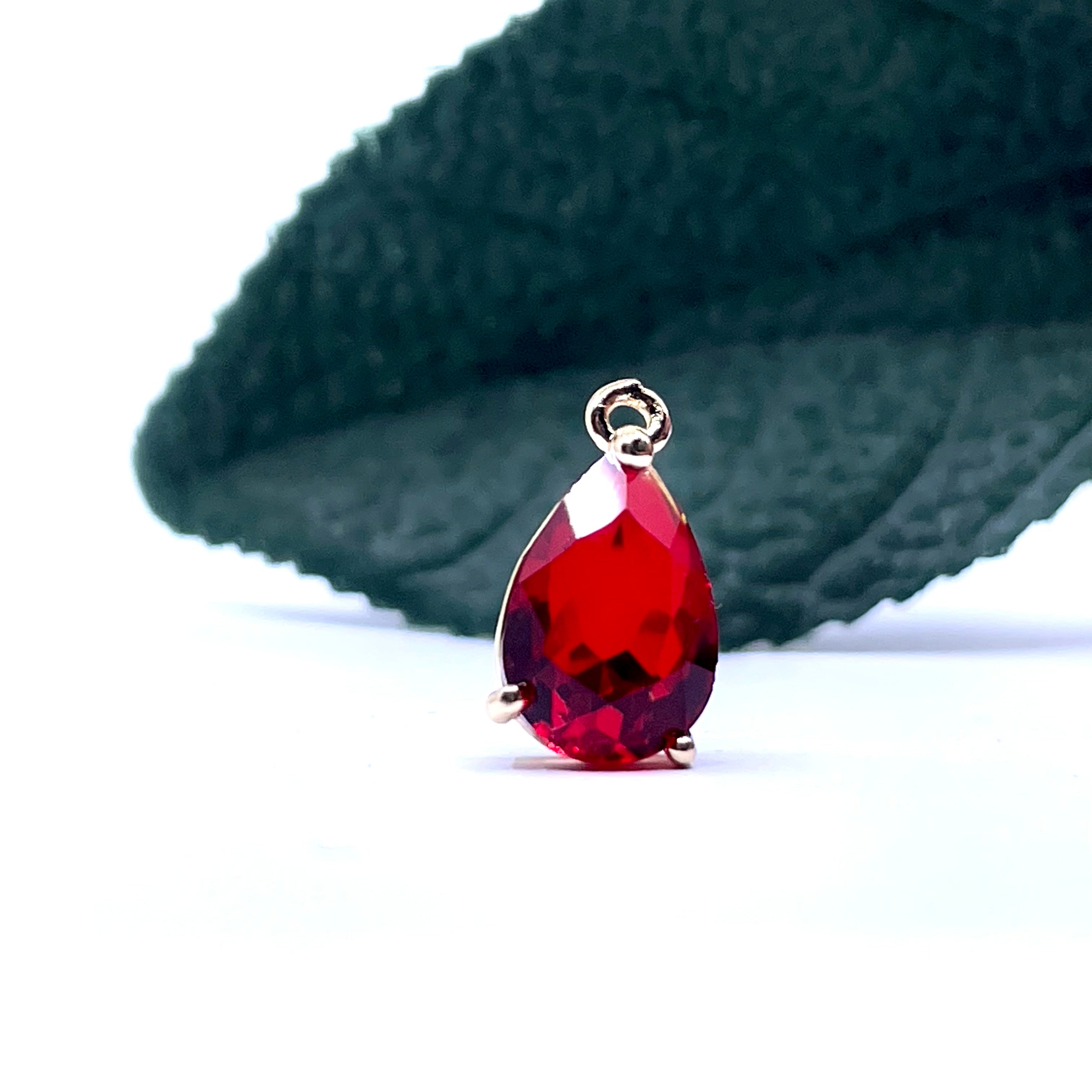 A faceted teardrop-shaped red gemstone pendant with a silver bail, displayed on a white surface with a dark green textured leaf in the background.
