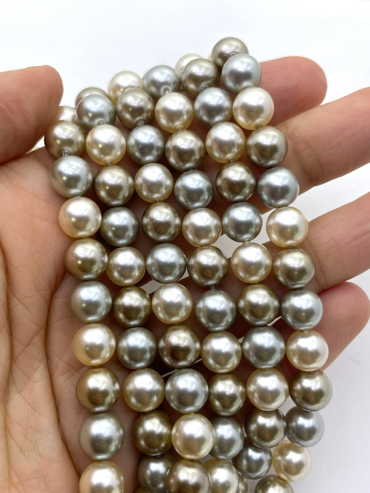 A close-up of a hand holding a strand of large, round shell pearls in a champagne to silver-gray color range against a plain white background.