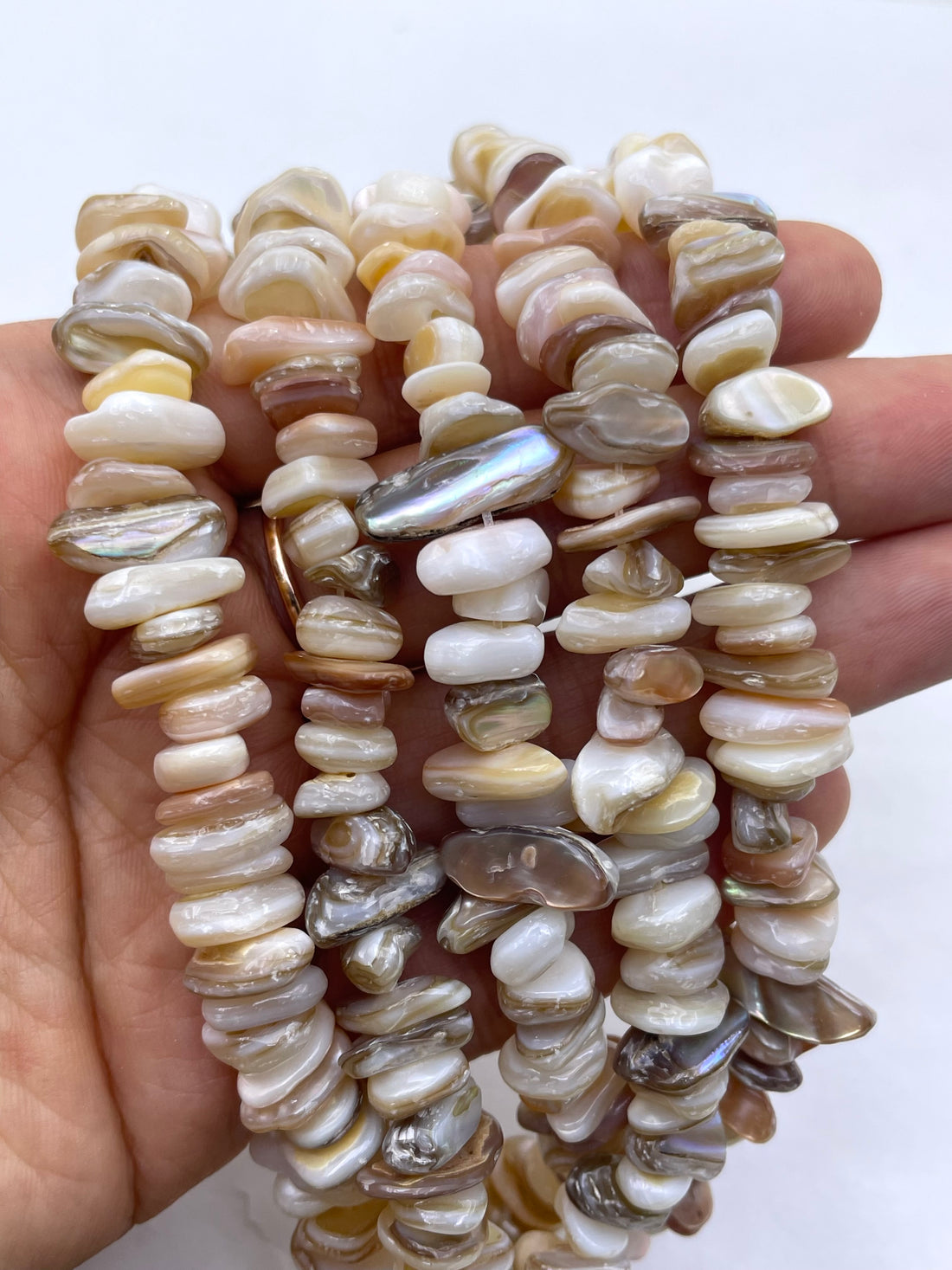 A close-up of a strand of natural stone chips or beads in mixed earthy tones including cream, beige, brown, gray, and translucent milky pieces, arranged horizontally on a plain light background.