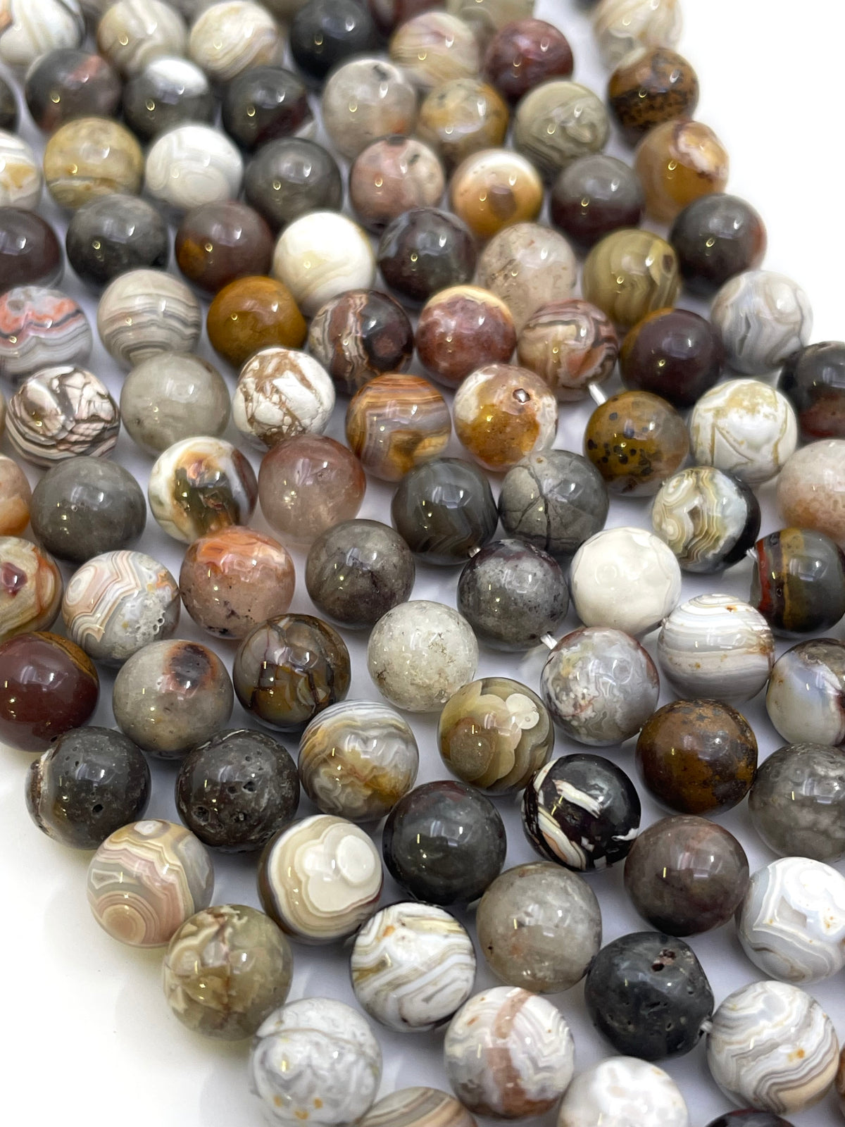A close-up of many polished round mexican crazy agate stone beads in mixed earthy colors and banded patterns, including gray, brown, white, black, and rust tones, arranged on a white background.