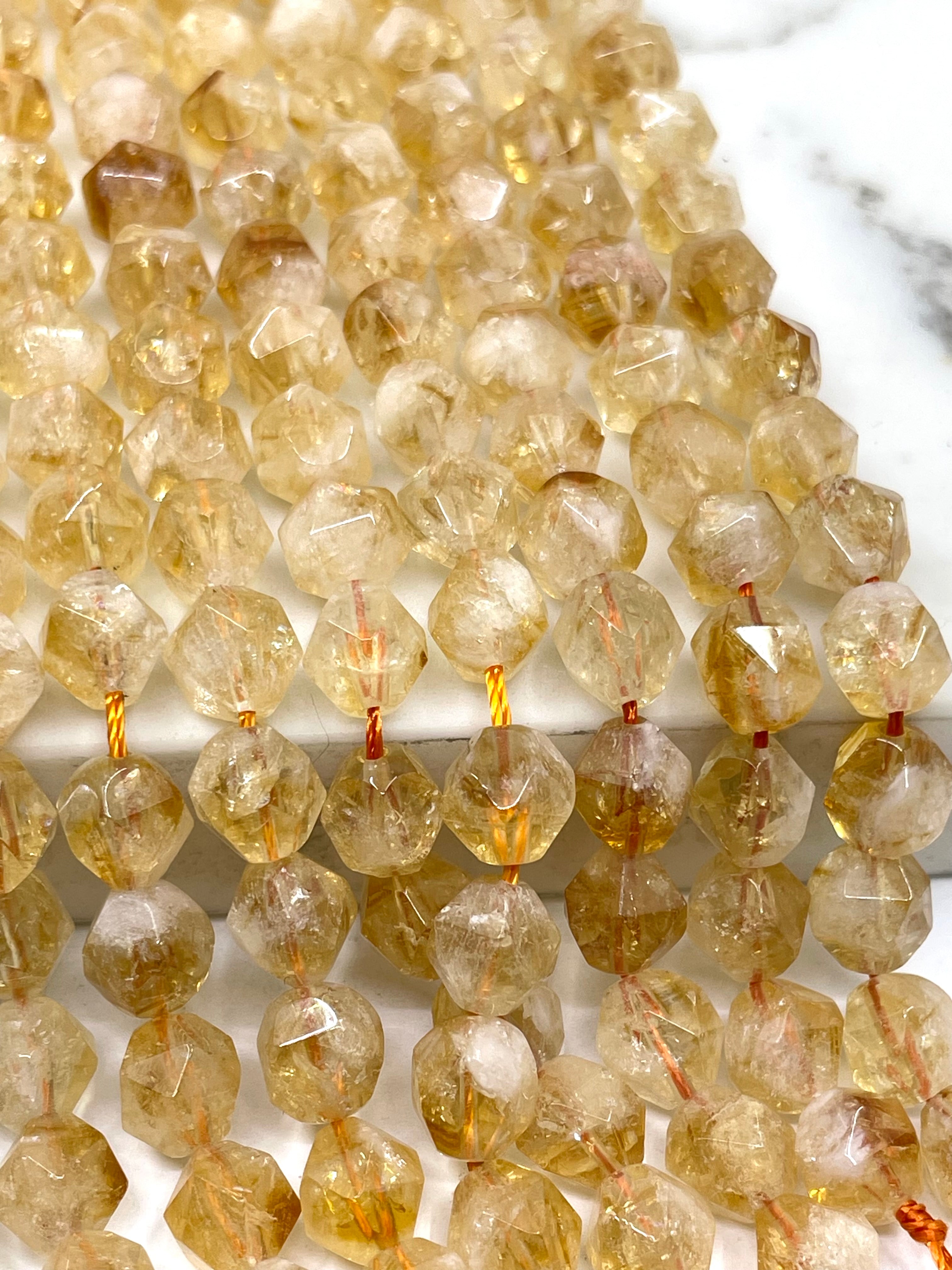 Faceted citrine beads in a light golden-yellow hue, strung on thin cords and arranged in parallel rows on a white marble surface.
