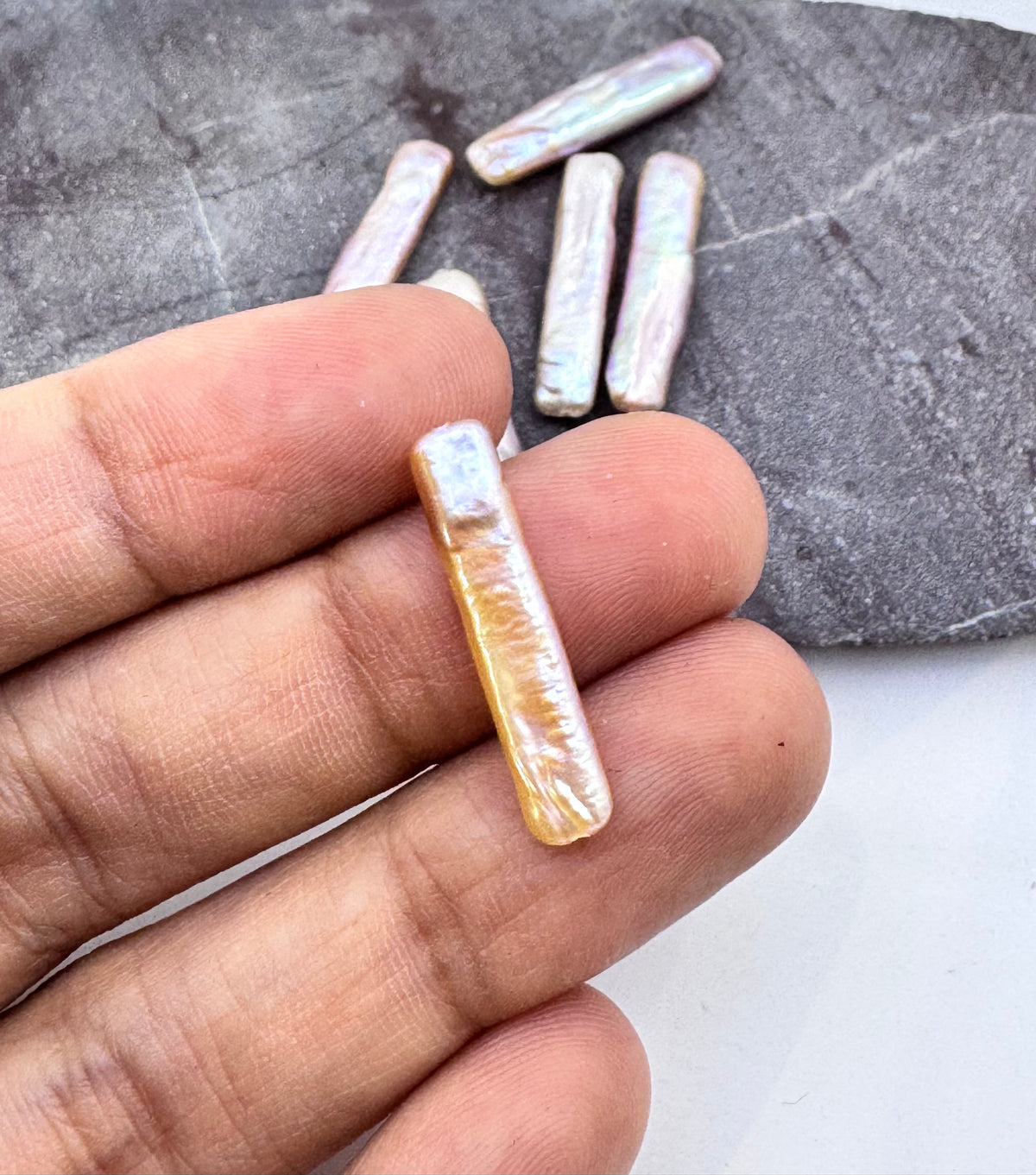 A hand holding a small, elongated iridescent shell bead, with several similar beads scattered on a dark textured surface in the background.