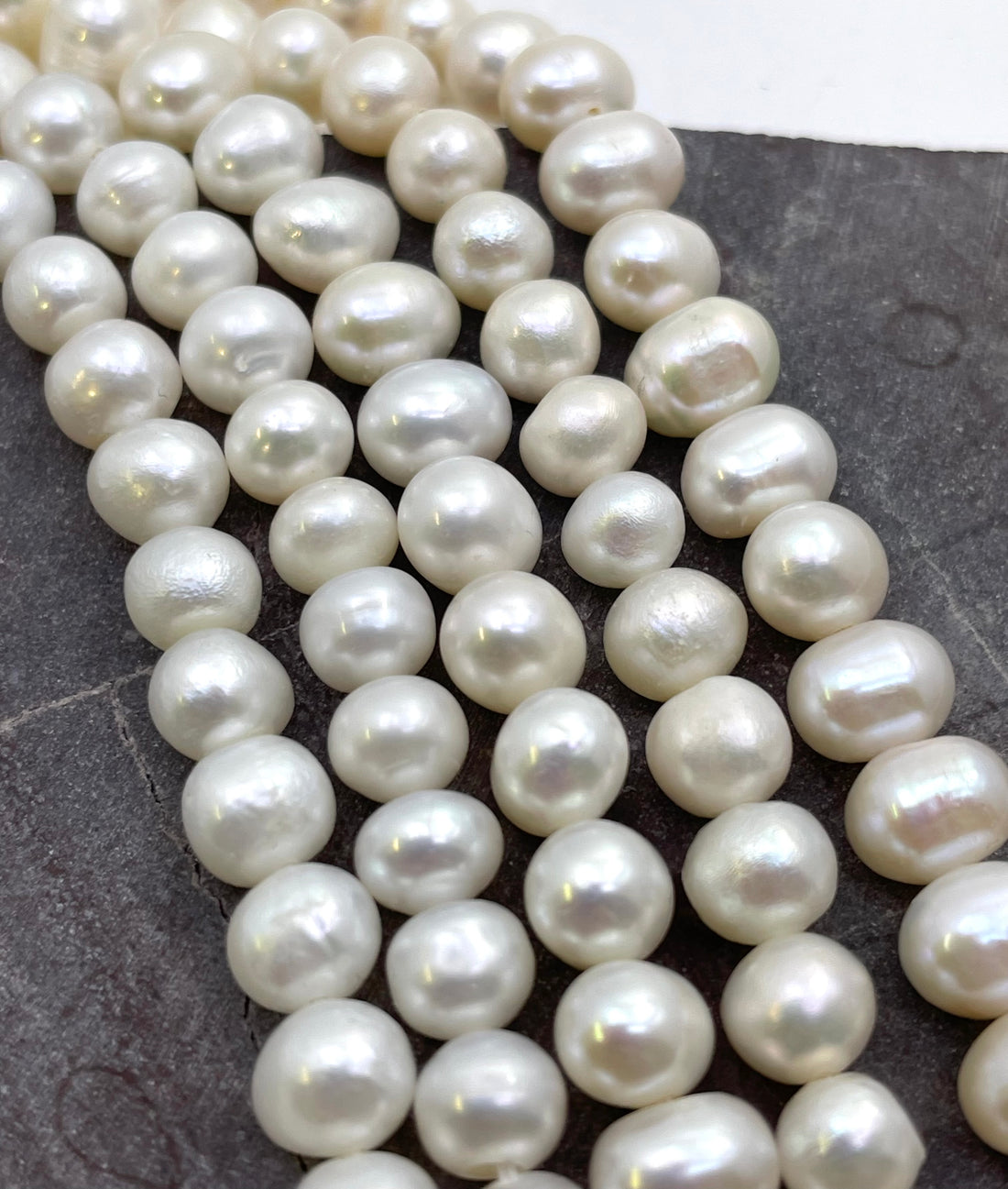 Close-up of multiple strands of irregular potato shape white freshwater pearls draped over a dark gray surface.