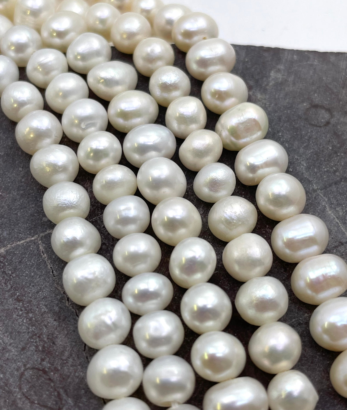 Close-up of strands of irregular white potato shape freshwater pearls arranged diagonally on a dark gray textured surface.