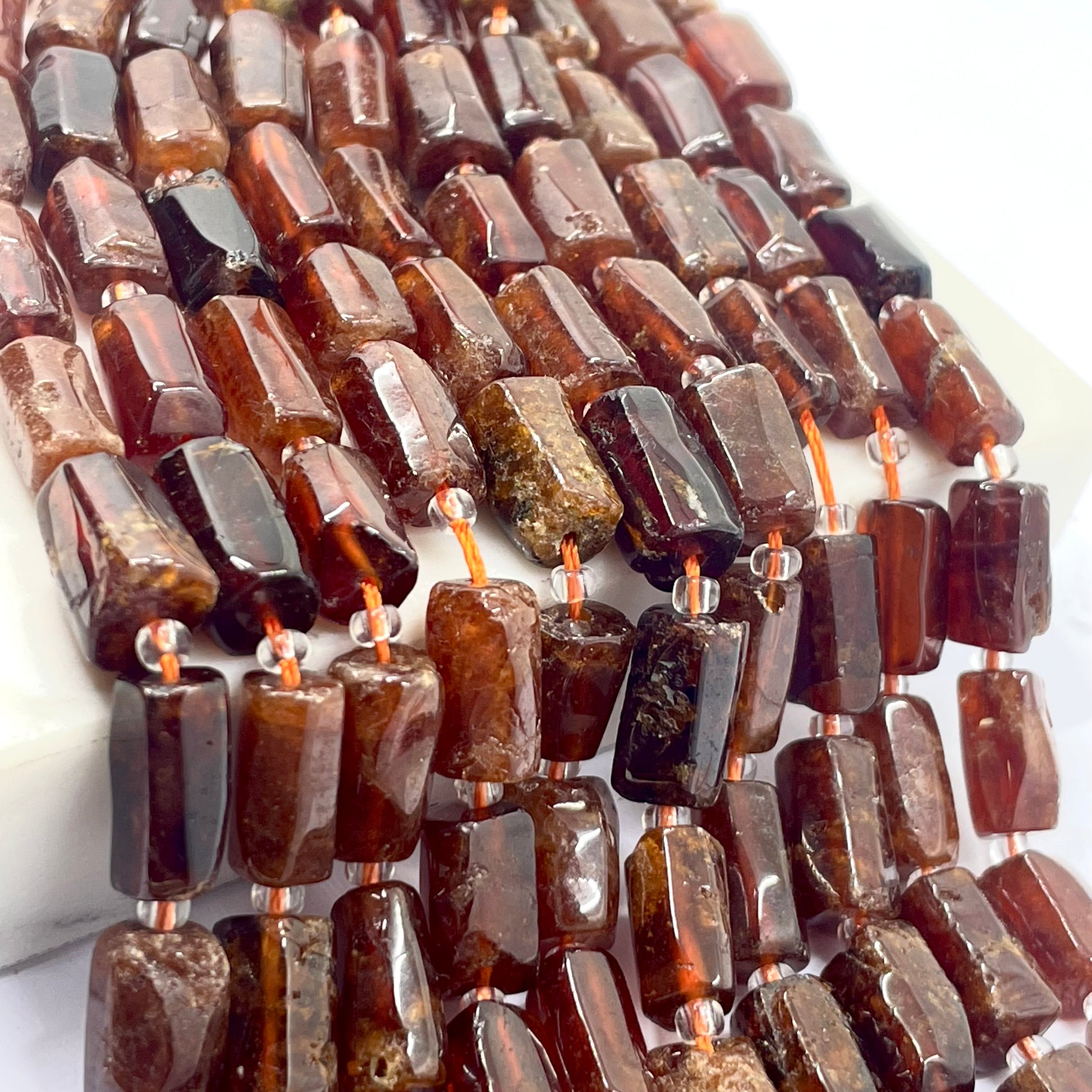 Close-up of multiple strands of polished brown translucent stone garnet beads with a faceted rectangular shape, strung on copper-colored cords over a white surface.