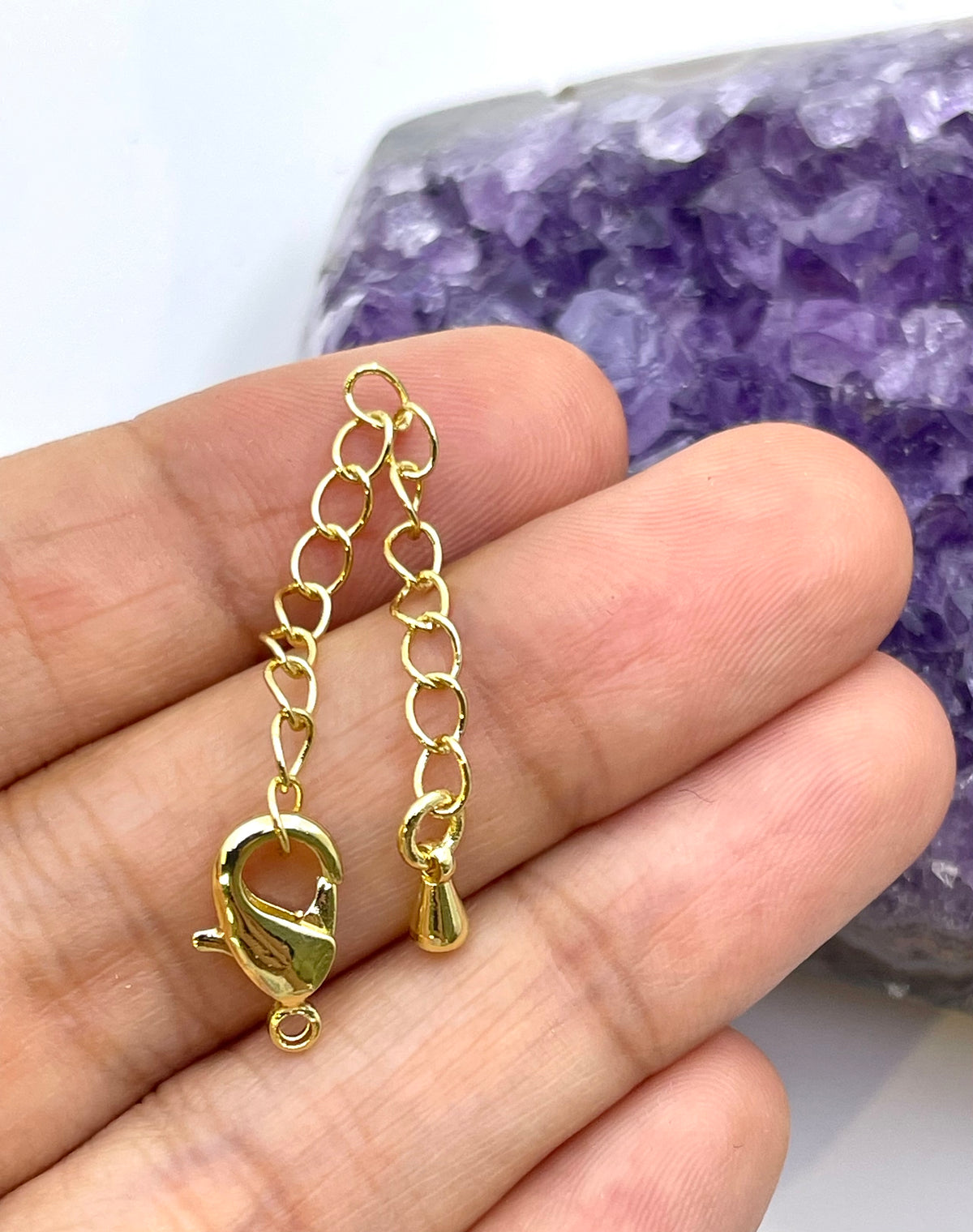 A close-up of a hand holding a small gold chain extender with a lobster clasp and a small bead, with a purple crystal geode in the background.