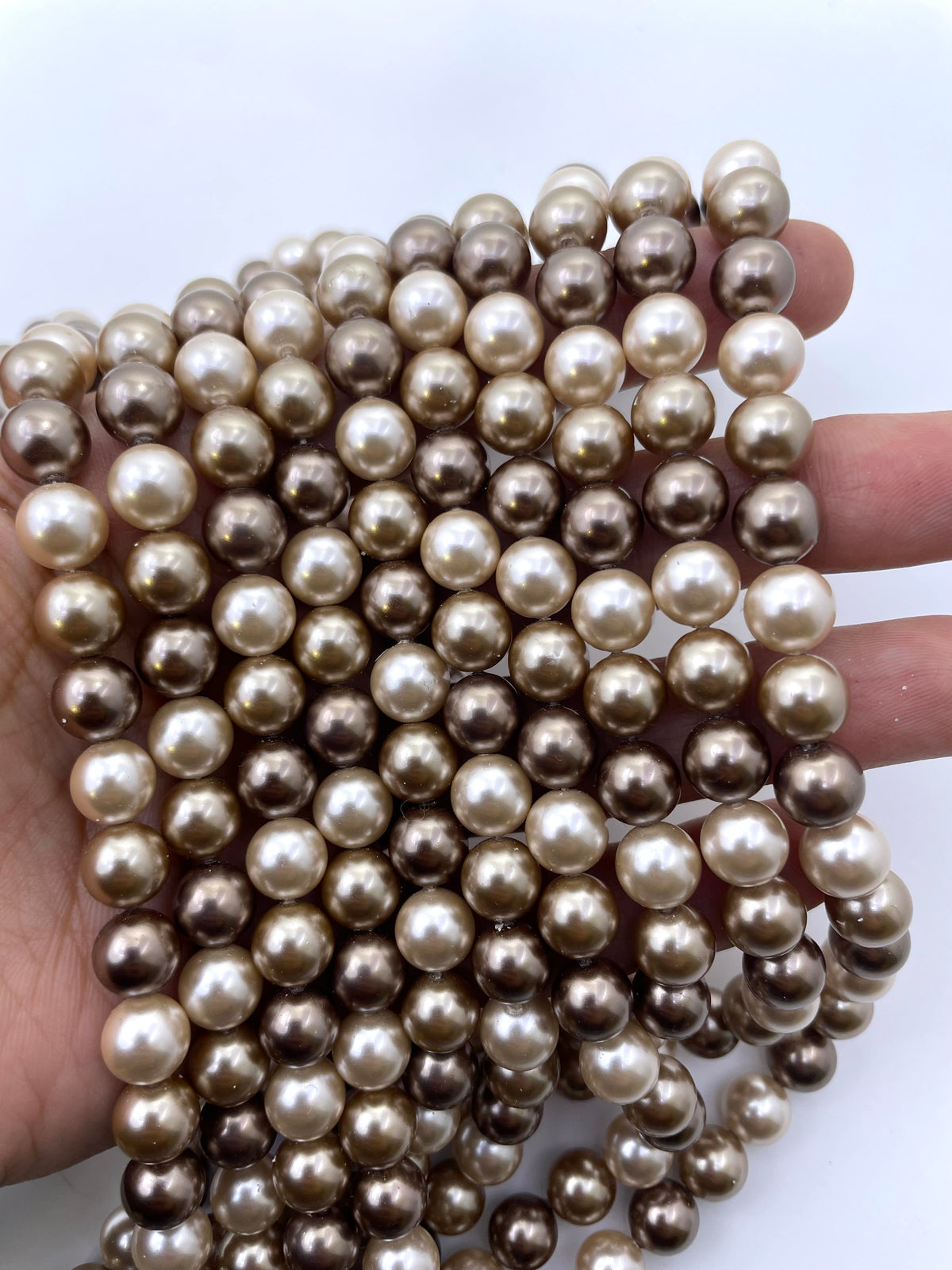 A hand holding a dense strand of round shell pearls in warm champagne, beige, and taupe tones against a plain white background.