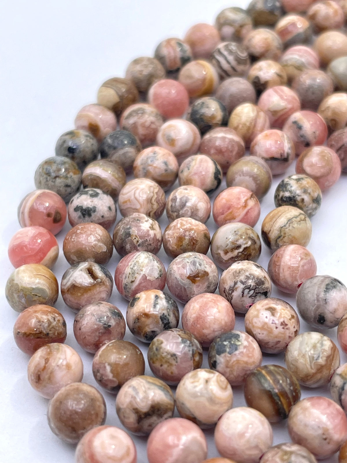 A close-up of polished round rhodochrosite gemstone beads in a marbled pattern of pink, beige, brown, and gray, arranged in a curved line on a white background.