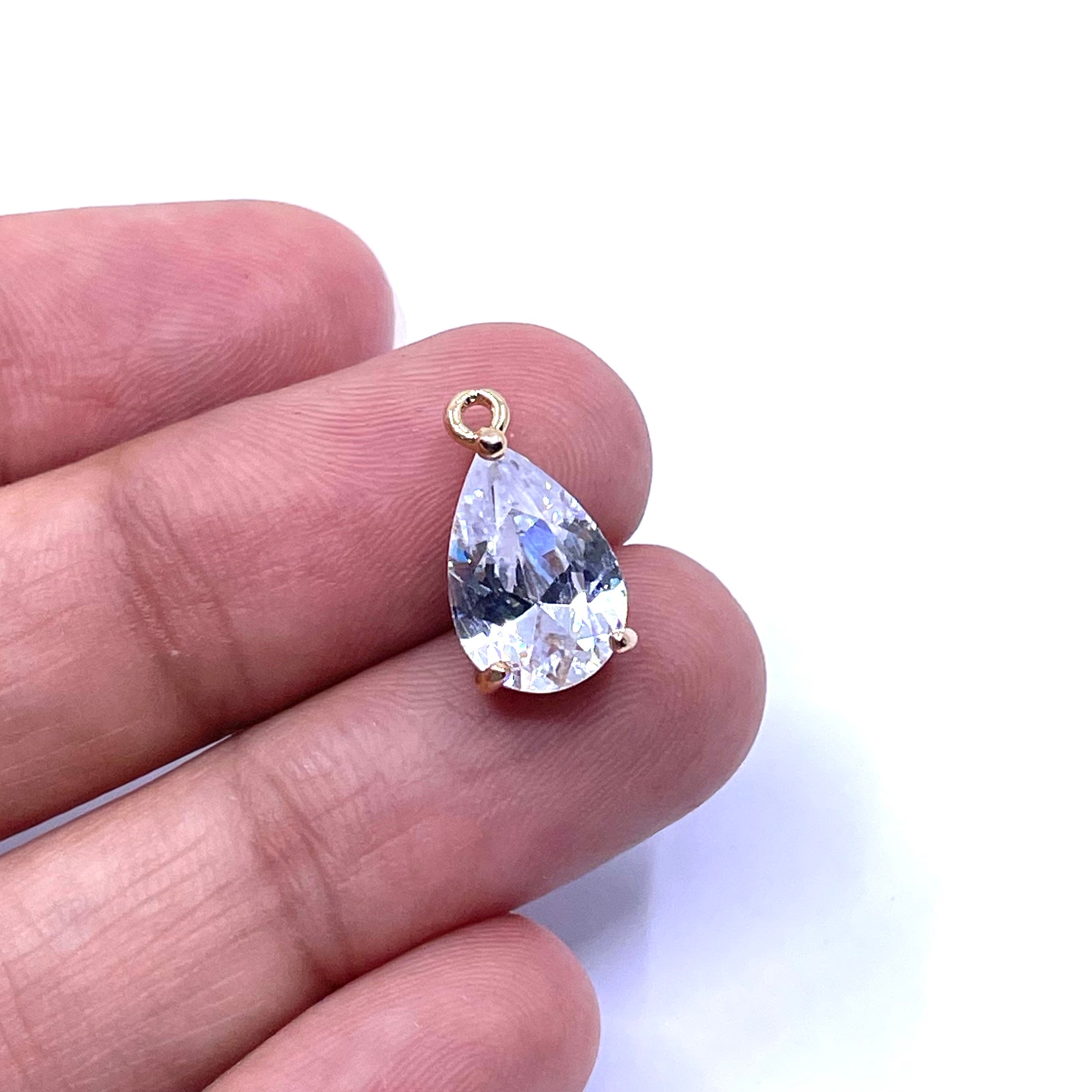 A teardrop-shaped clear gemstone pendant in a gold prong setting, resting on a person's fingers against a white background.