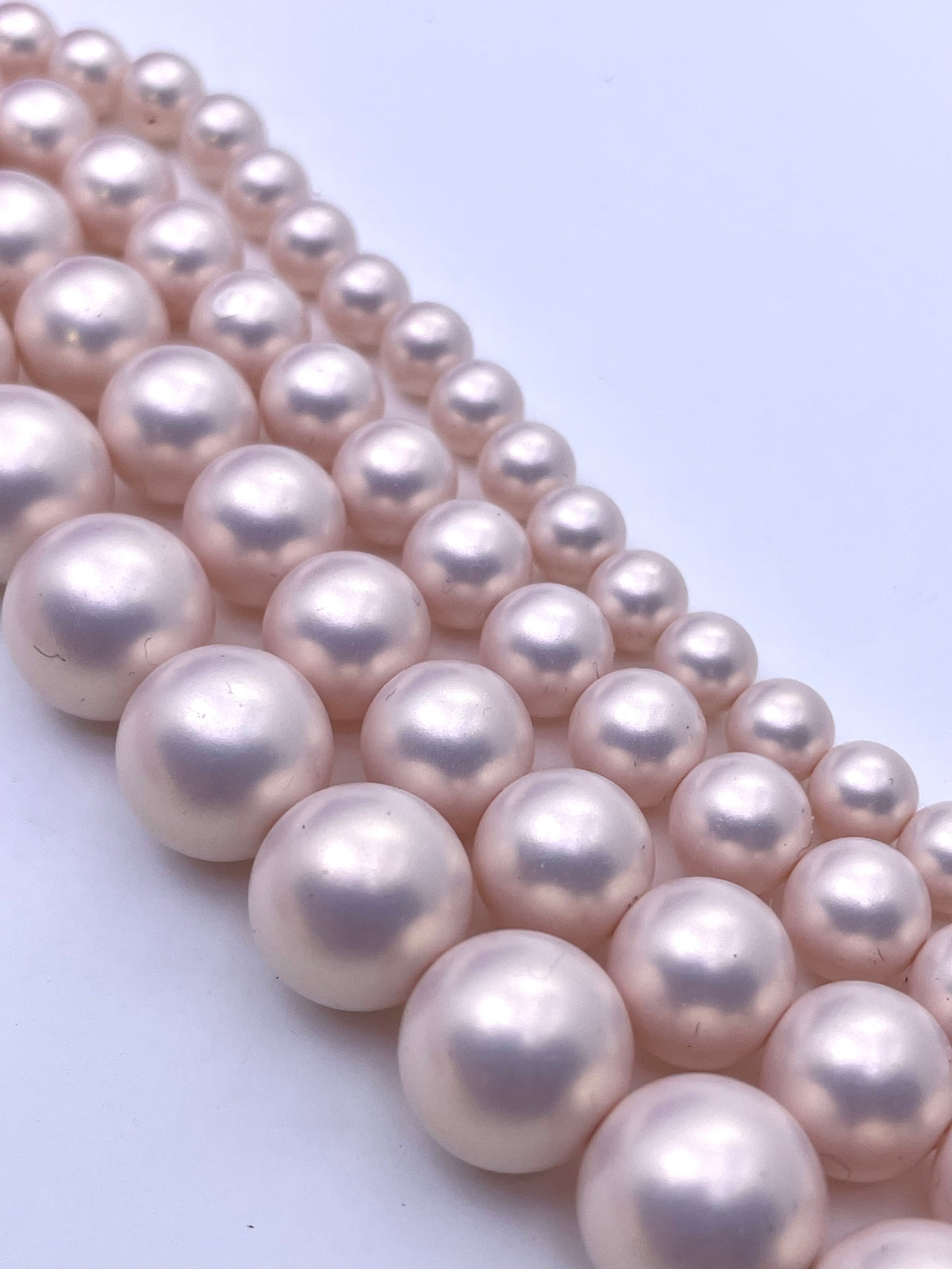 Multiple strands of round shell pearl beads in a light pinkish-white lustrous finish, arranged diagonally on a plain light background.
