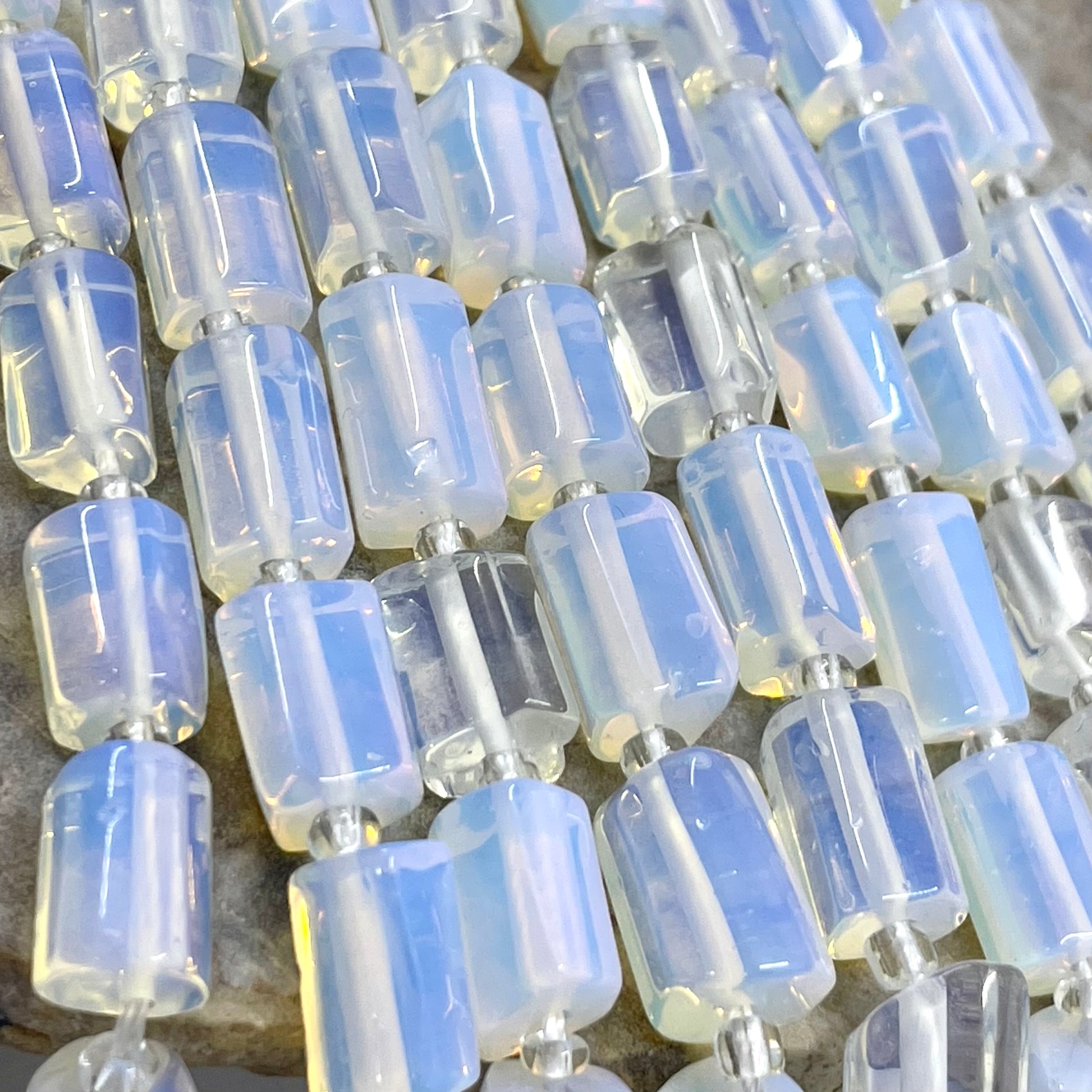 A close-up of a strand of translucent light blue and white opalite cylindrical beads with small silver spacer rings, laid out on a neutral surface.