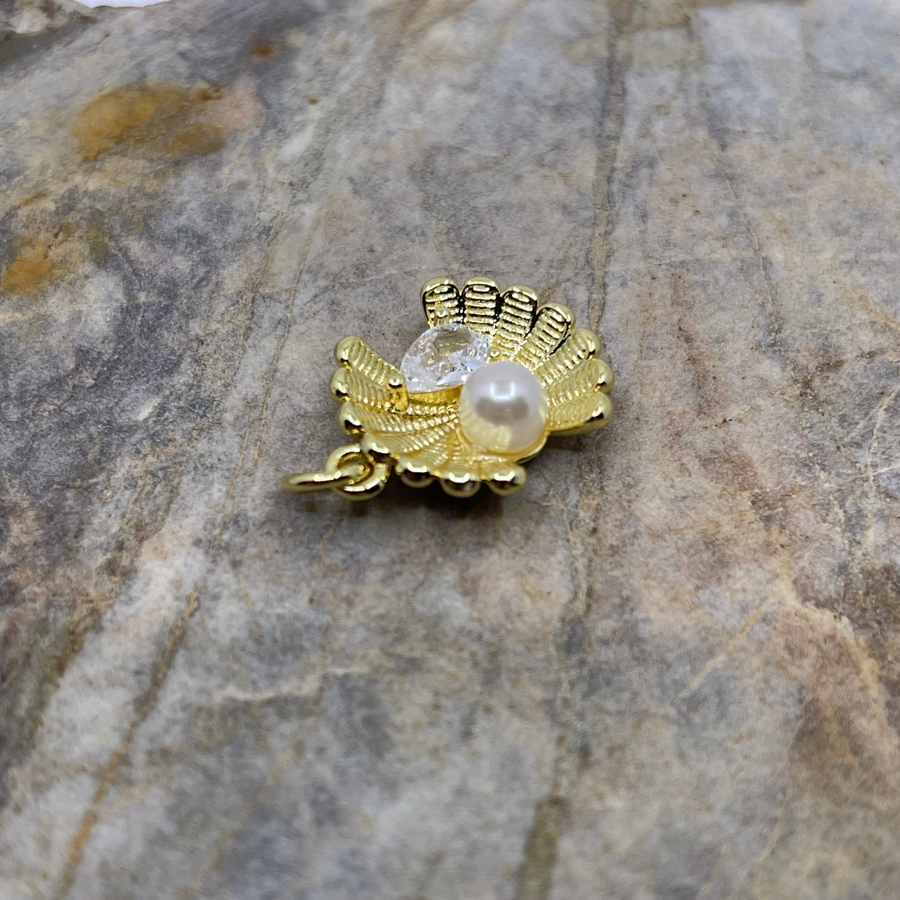 A gold-toned shell-shaped pendant charm featuring a central clear faceted crystal and a white pearl, resting on a textured gray stone surface.