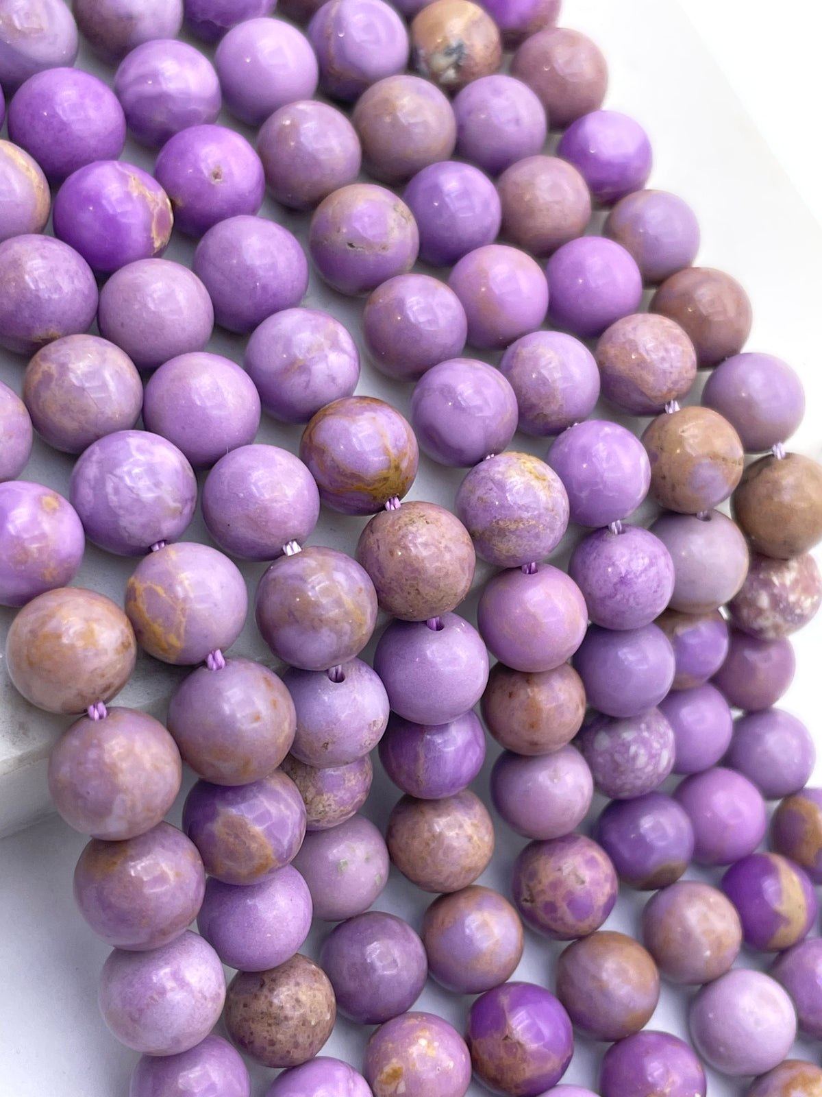 Purple and lavender round phosphosiderite gemstone beads with natural marbled patterns, arranged in a diagonal line on a white background.