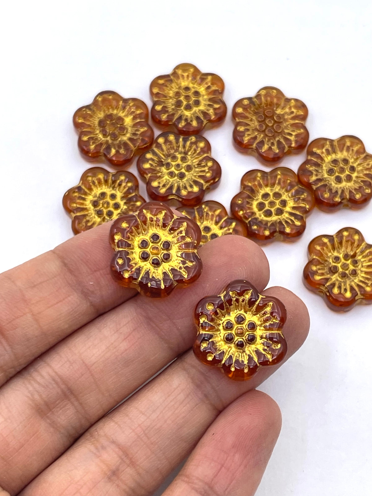 Several amber-brown translucent flower-shaped beads with gold glitter centers are displayed on a white background, with two larger beads resting on a person's fingers to show scale.