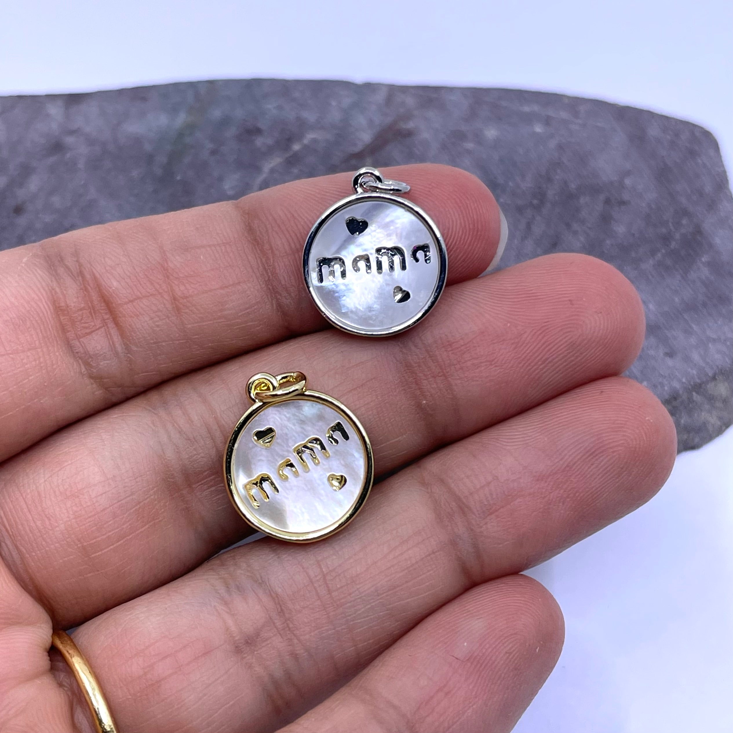 Two round pendant charms engraved with 'mama' and small heart shapes, one with a silver-toned bezel and one with a gold-toned bezel, held on a person's fingers against a gray stone background.