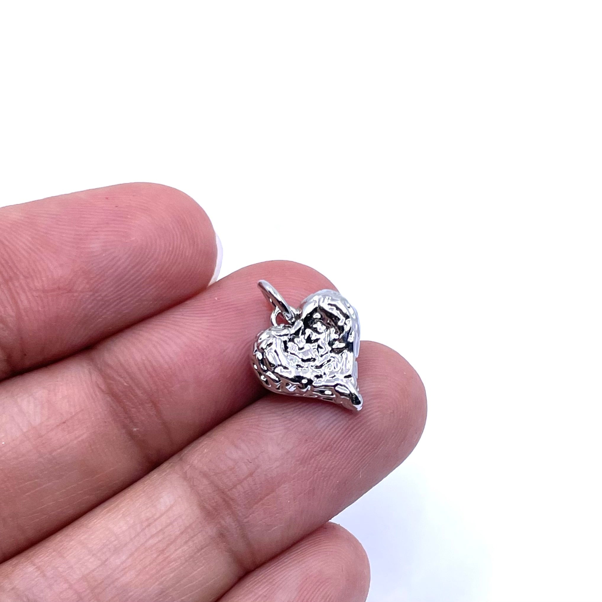 A small textured silver heart charm held between two fingers against a plain white background.