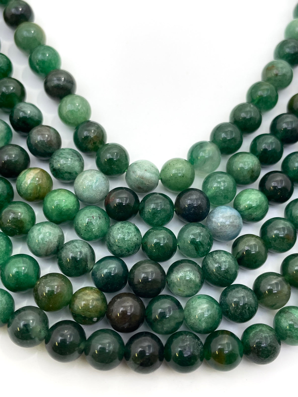 Multiple strands of polished round green lepidolite gemstone beads, varying from light forest green to dark emerald with natural inclusions, arranged on a plain white background.