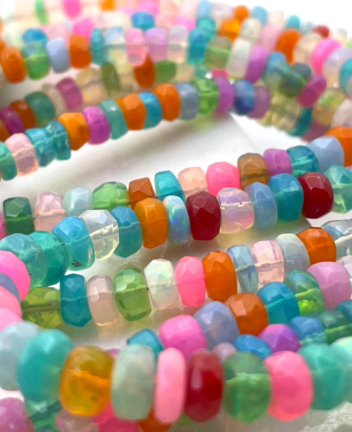 Close-up of multiple strands of faceted disc ethopian opal beads in assorted bright colors including pink, teal, orange, green, red, yellow, and clear, arranged on a white background.