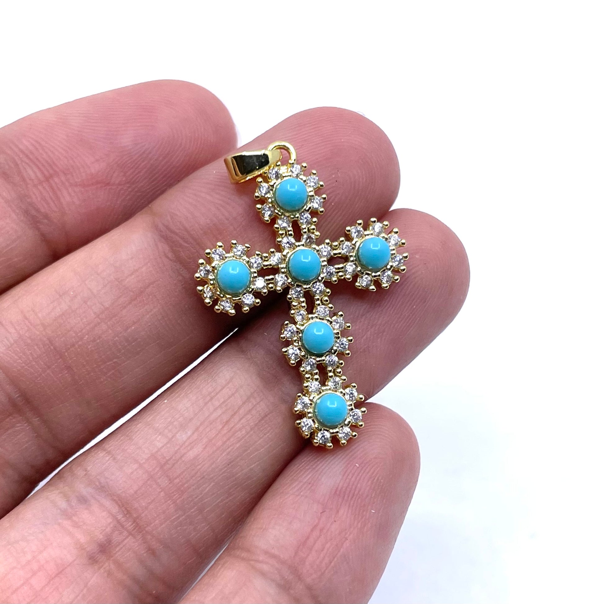 A gold cross pendant with SIX light blue turquoise-like round stones, held on a person's fingers against a plain white background.