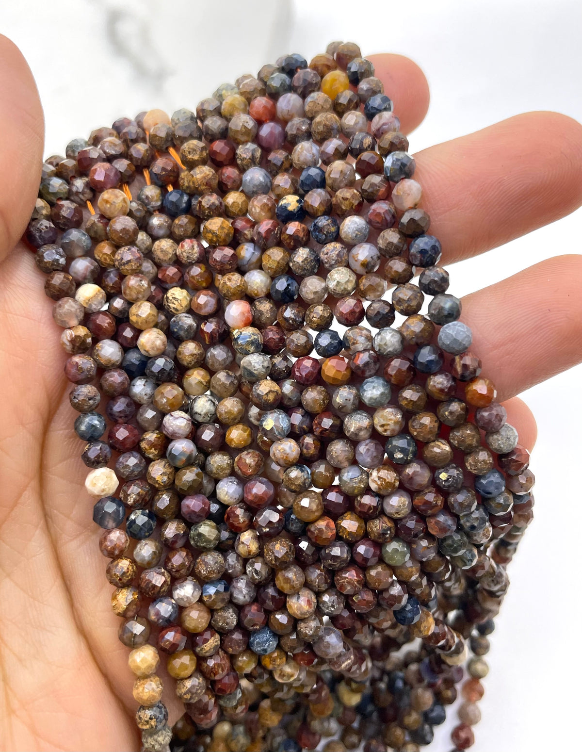 A close-up of a hand holding a bundle of small faceted pietersite gemstone beads in mixed earthy and metallic colors.