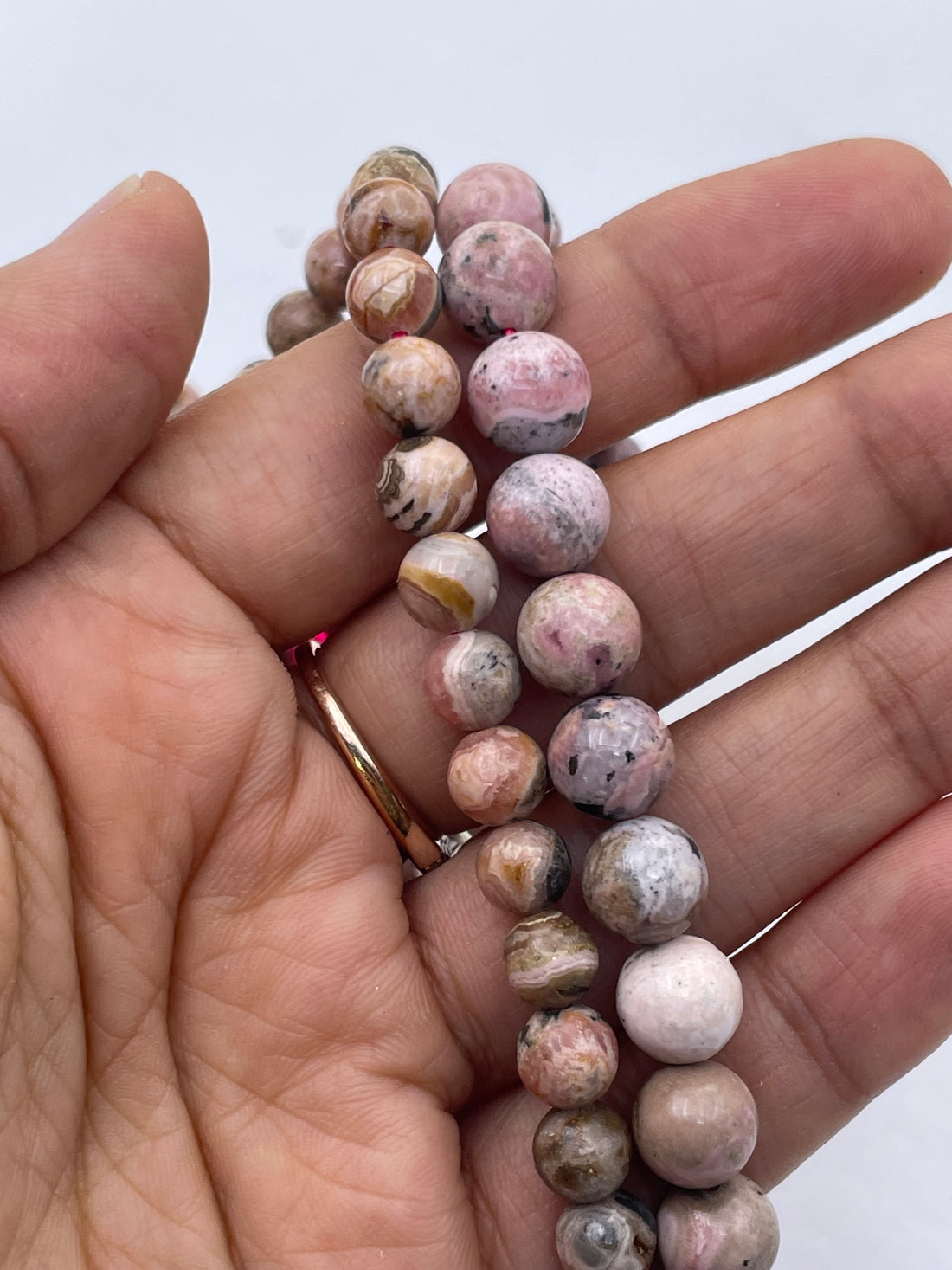 A hand holding a bundle of round, polished pink and brown rhodochrosite stone beads with natural marbled patterns, set against a plain light gray background.