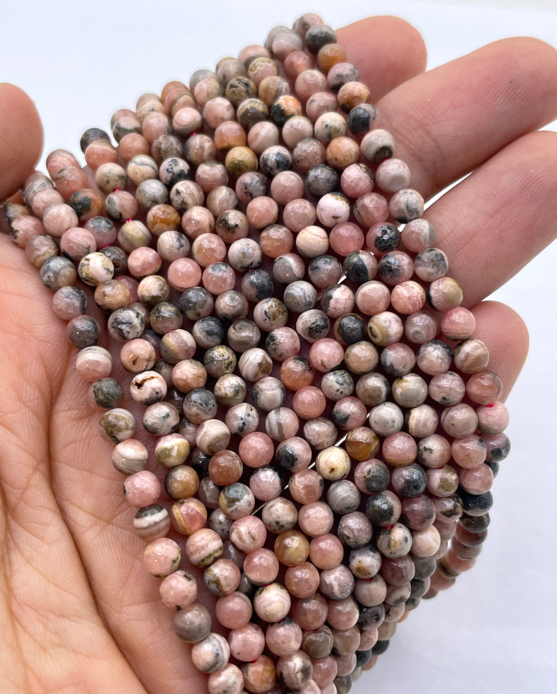 Curved strands of faceted round rhodochrosite beads made from pinkish stone with natural marbling, speckling, and dark inclusions, photographed on a white background.