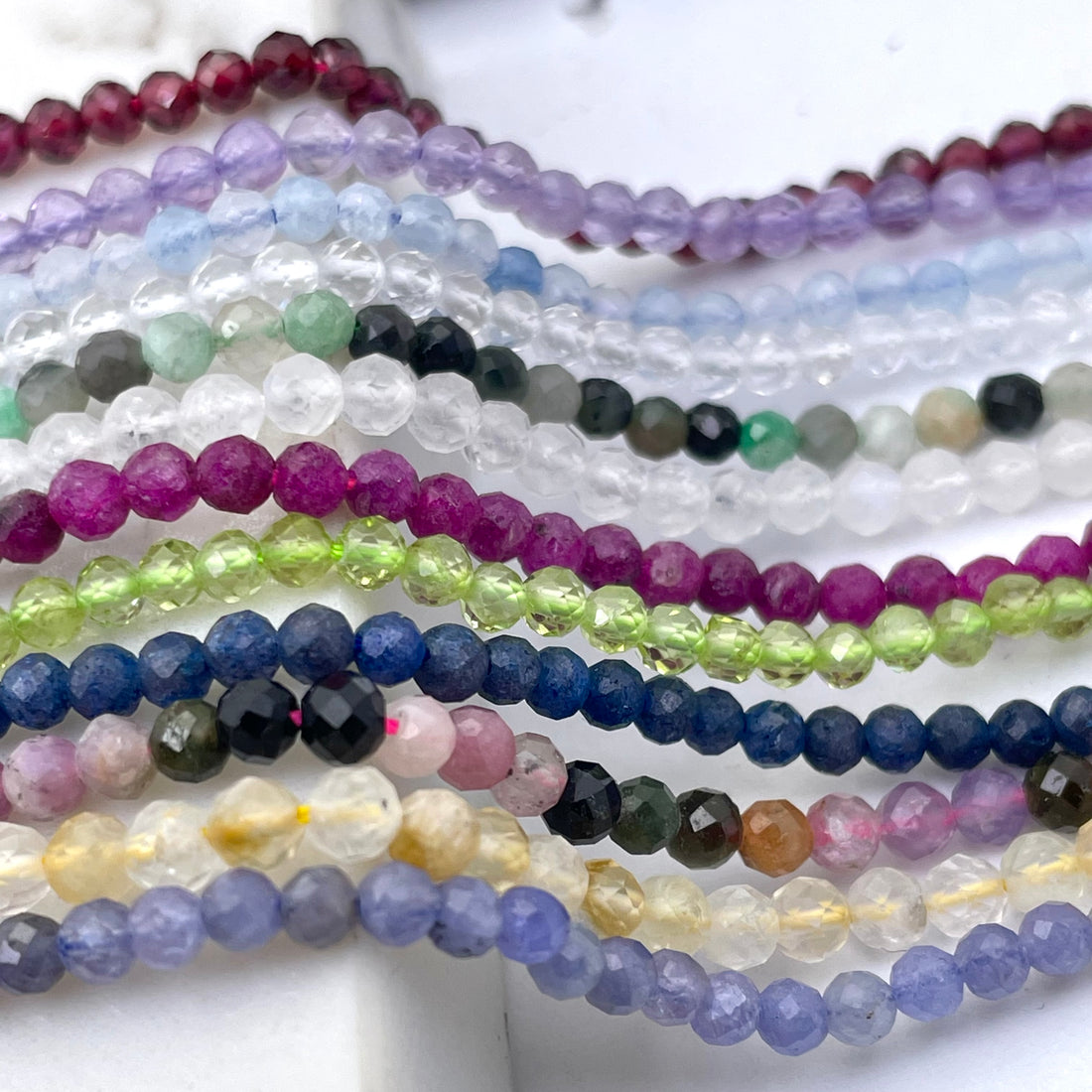 Multiple strands of faceted round gemstone beads in assorted colors, including lavender, yellow, blue, green, pink, purple, gray, and red, arranged vertically on a white background.