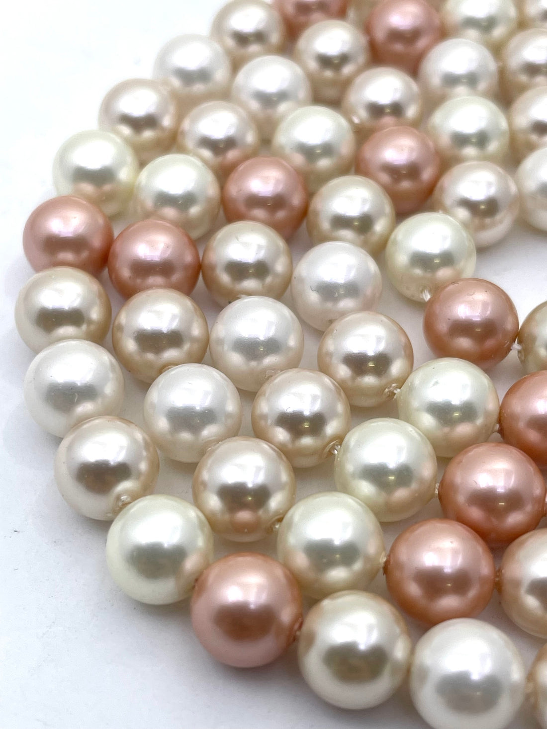 Close-up of strands of round shell pearls in white, cream, and soft peach-pink tones on a light gray background.