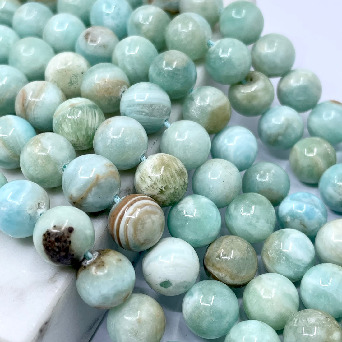 A close-up of round light blue-green hemimorphite gemstone beads with natural marbling and one striped accent bead, arranged on a white marble surface.